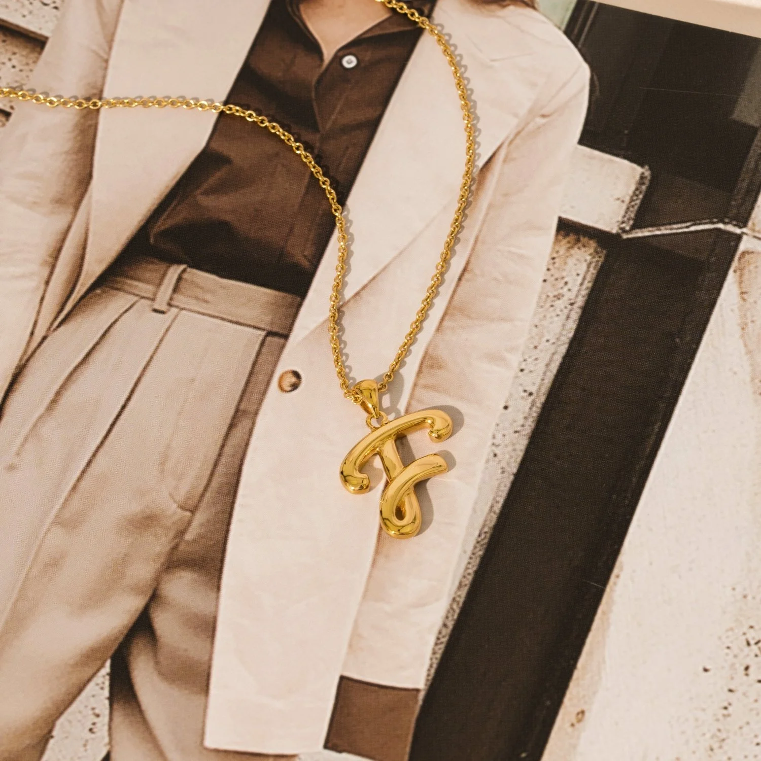 Close-up of a gold necklace with a large gold pendant, worn over a beige blazer, black shirt, and beige trousers, against a background featuring a black-and-white patterned object.