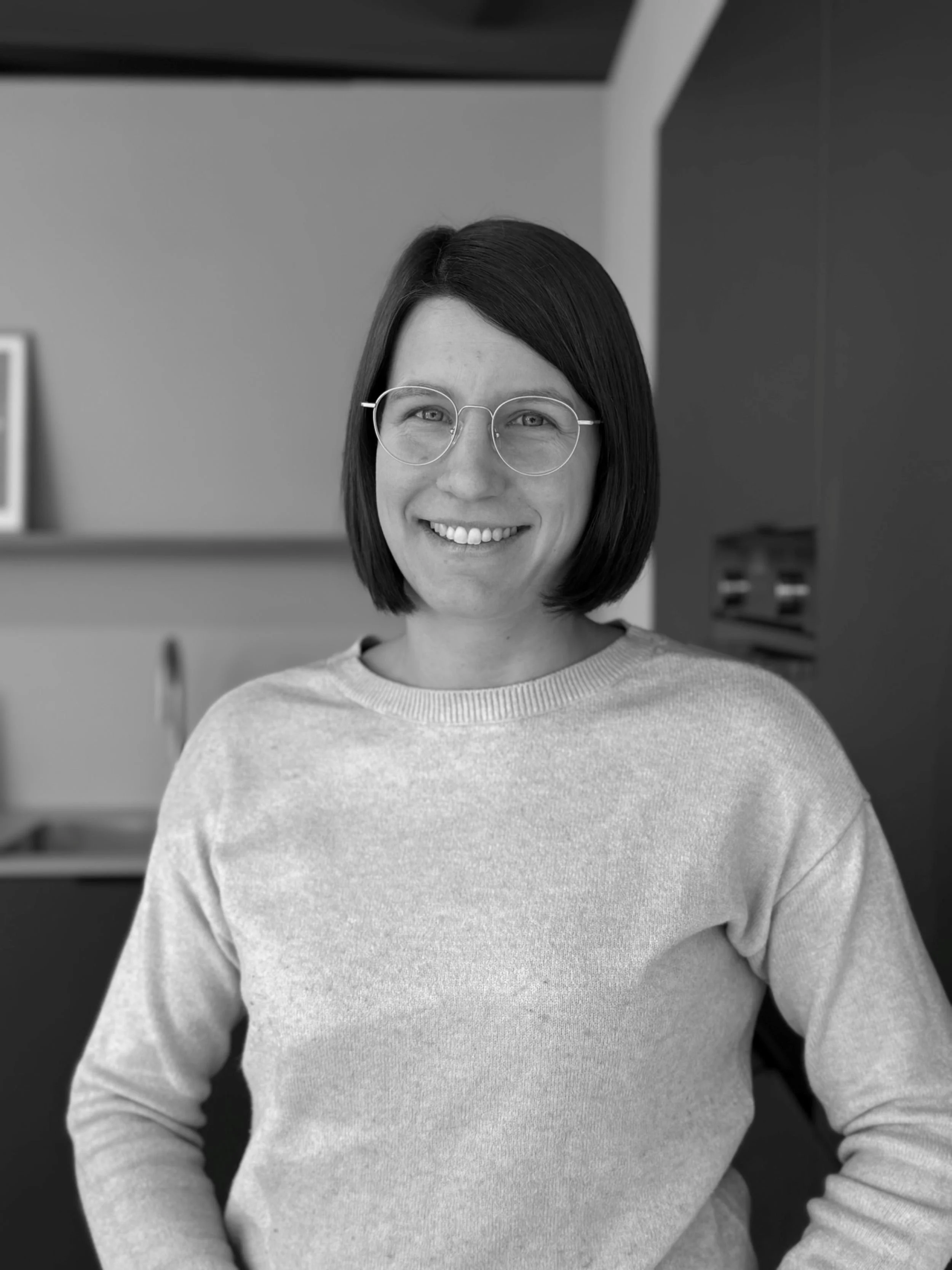 Schwarz-weiß-Fotografie einer lachenden jungen Frau mit kurz schwarzem Haar und Brille, im Hintergrund eine moderne Innenarchitektur.