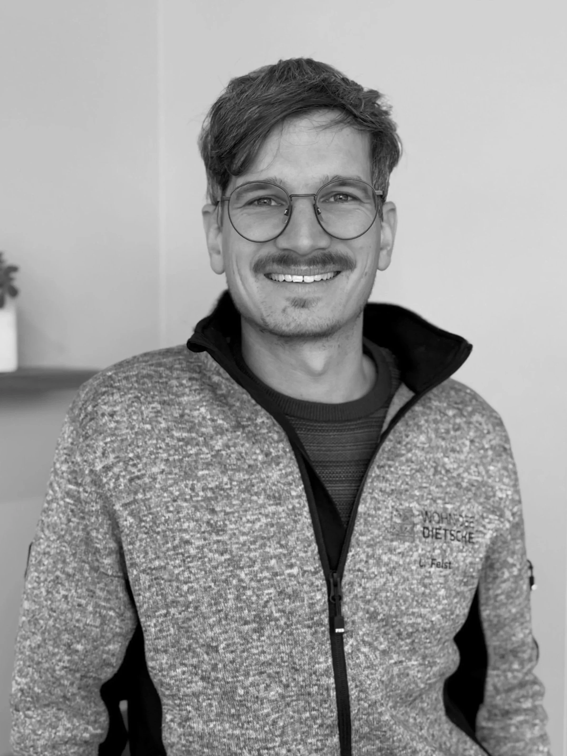 Schwarz-weißer Porträt eines lachenden Mannes mit Brille, kurzen dunklen Haaren, Bart und Trummeln, trägt einen Zipp-Up-Sweater, im Hintergrund eine weiße Wand und eine kleine Pflanze auf einer Ablage.