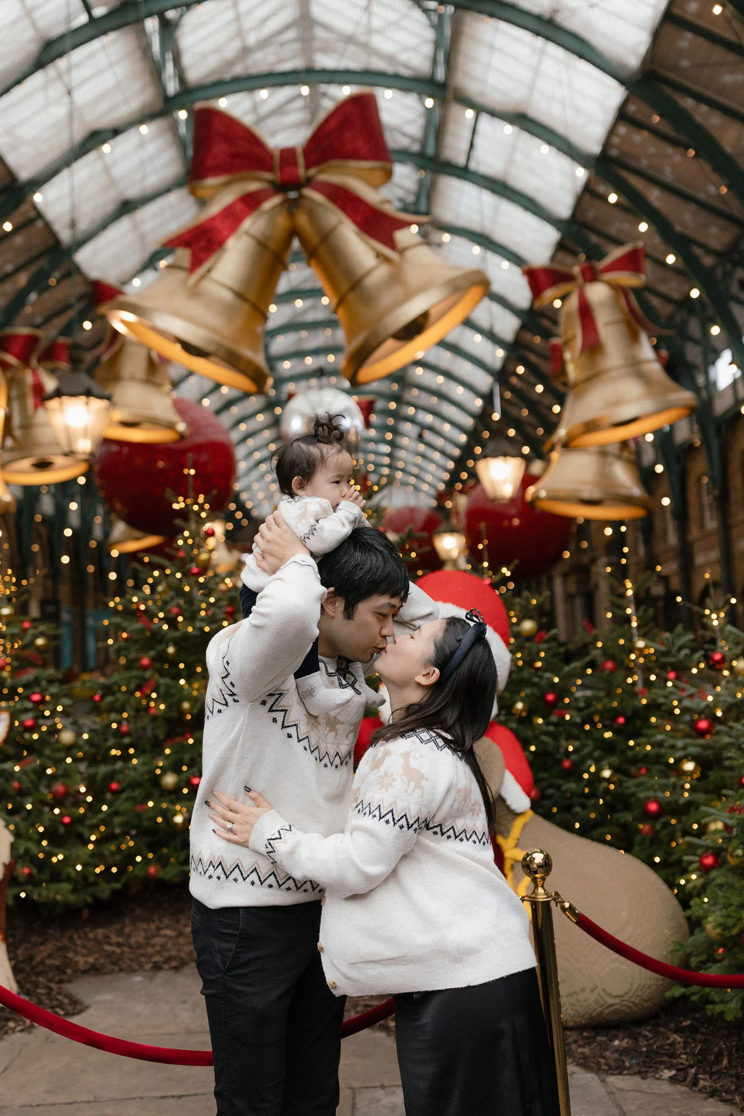 A family of three, parents and a young child, sharing a kiss in front of Christmas decorations, including a decorated Christmas tree, large gold bells with red bows, and hanging Christmas ornaments in an indoor setting with a high arched glass ceilin