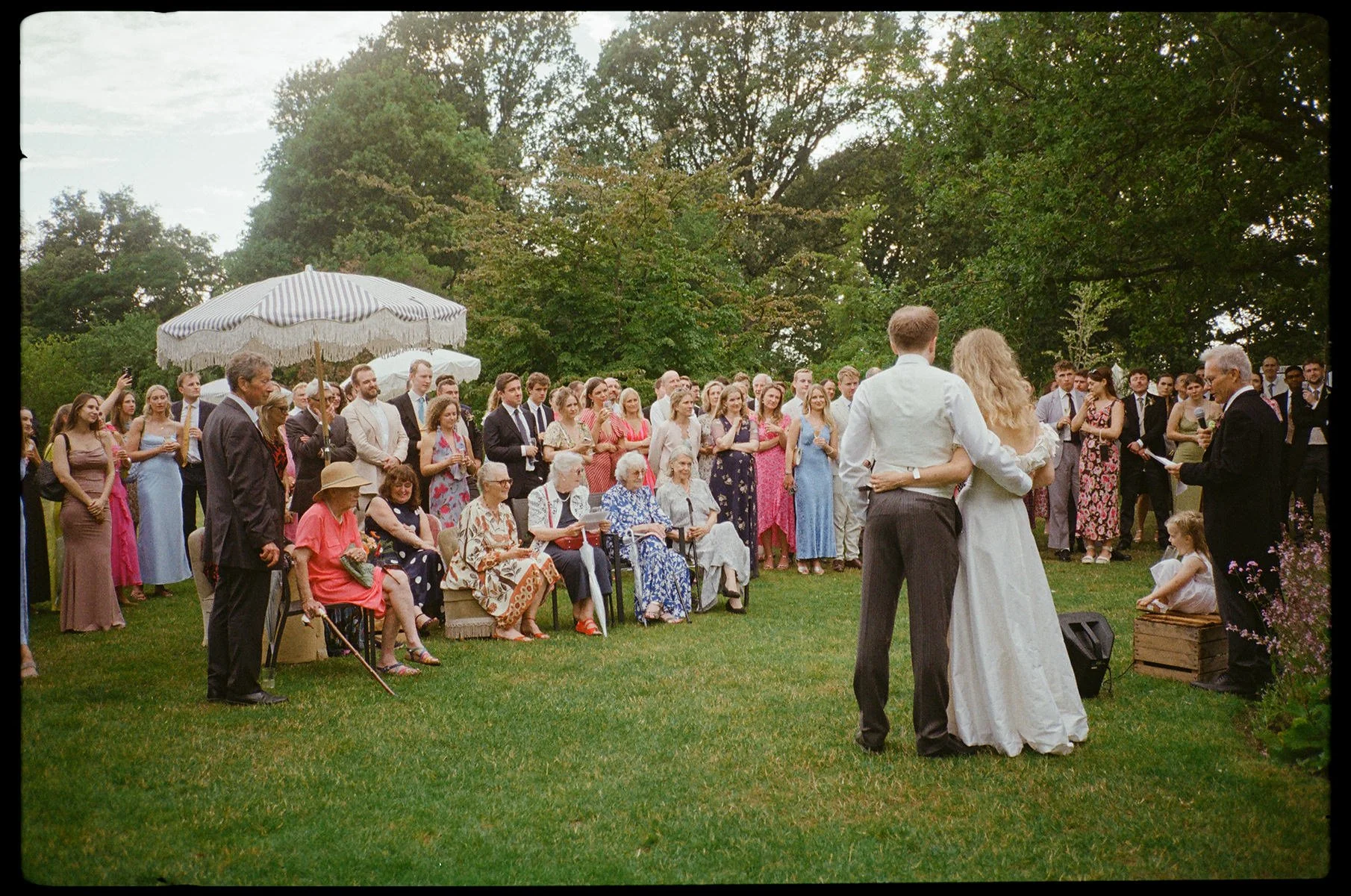 A wedding ceremony taking place outdoors on a lush green lawn, with a bride and groom standing close together in front of an officiant, and guests watching the ceremony, some seated and some standing, under umbrellas and surrounded by trees.
