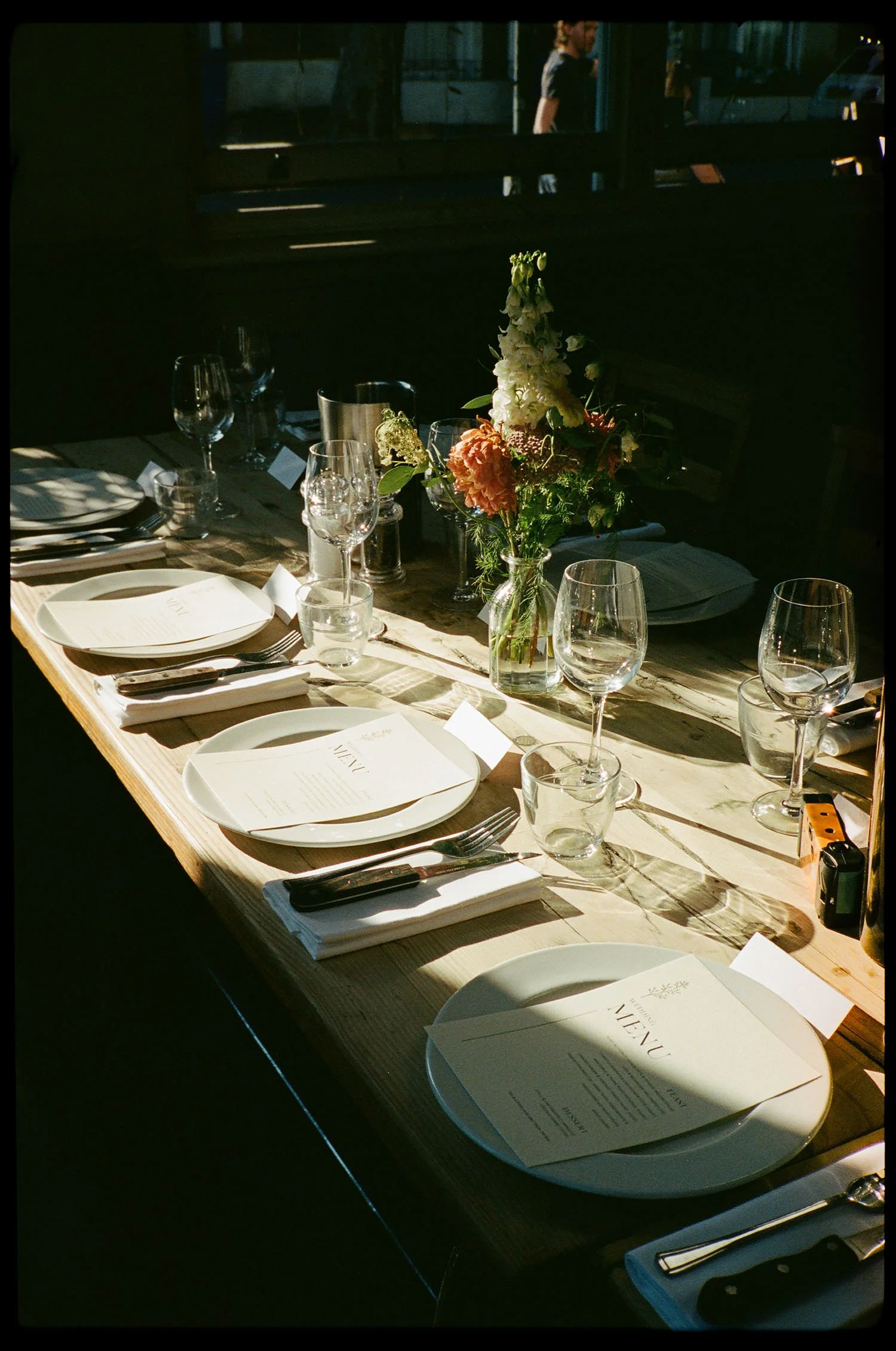 A table set for a meal with wine glasses, plates, utensils, and a printed menu. There is a floral centerpiece on the table and sunlight casting shadows on the table surface.