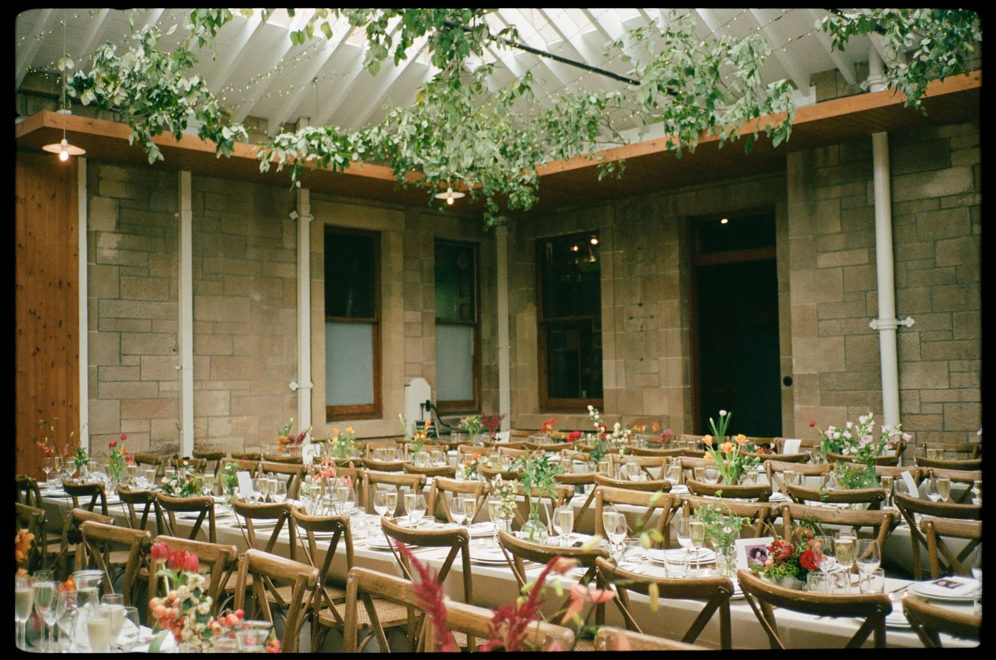 Indoor wedding reception setup with long tables decorated with flowers, surrounded by wooden chairs, in a rustic venue with brick walls, large windows, and greenery hanging from the ceiling with string lights.