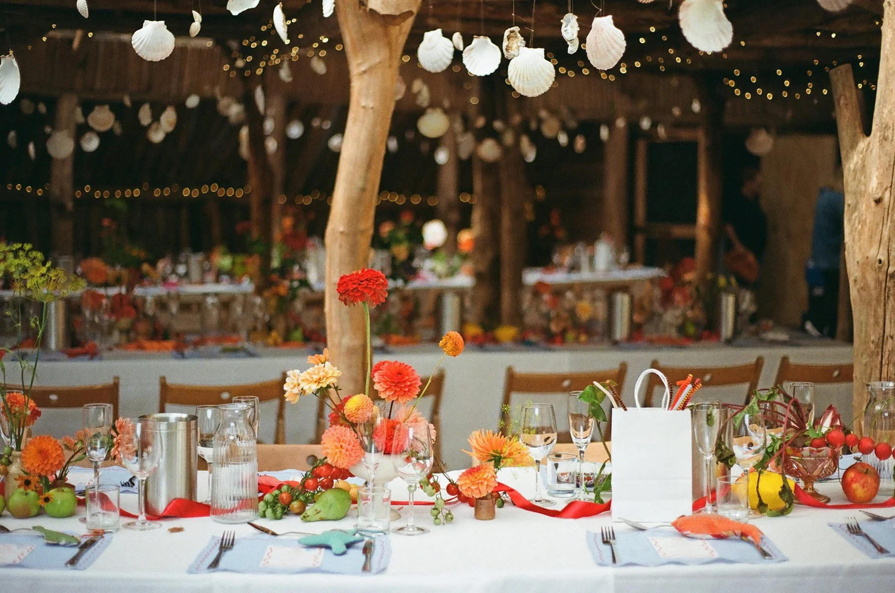 Decorated banquet table with colorful flowers, glassware, and table settings at an event in a rustic barn with hanging shell ornaments and fairy lights