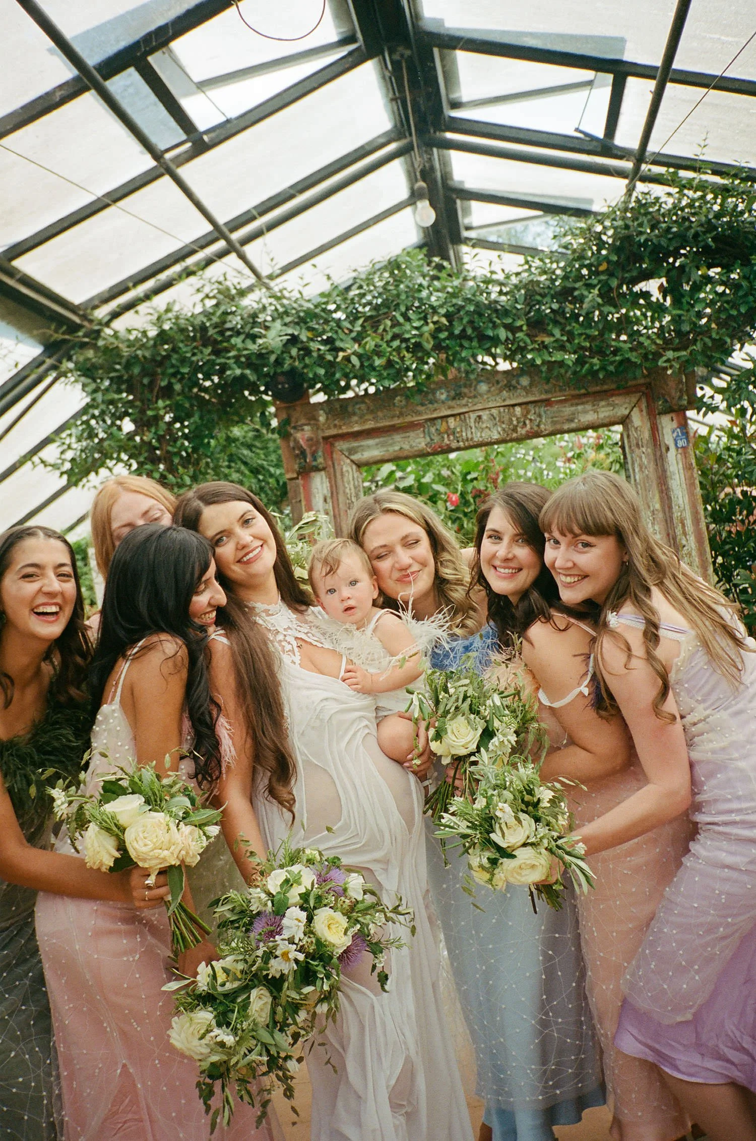 Group of women in pastel dresses holding bouquets of flowers, gathered around a bride, in a greenhouse  setting with greenery and a wooden frame in the background, celebrating a wedding.