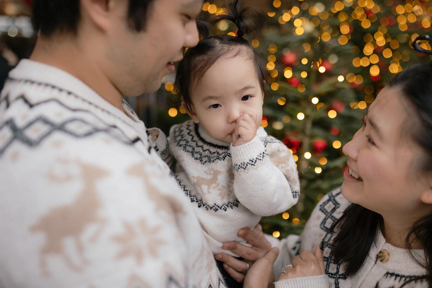 A man, woman, and little girl celebrate Christmas with a decorated tree in the background, all wearing matching holiday sweaters.