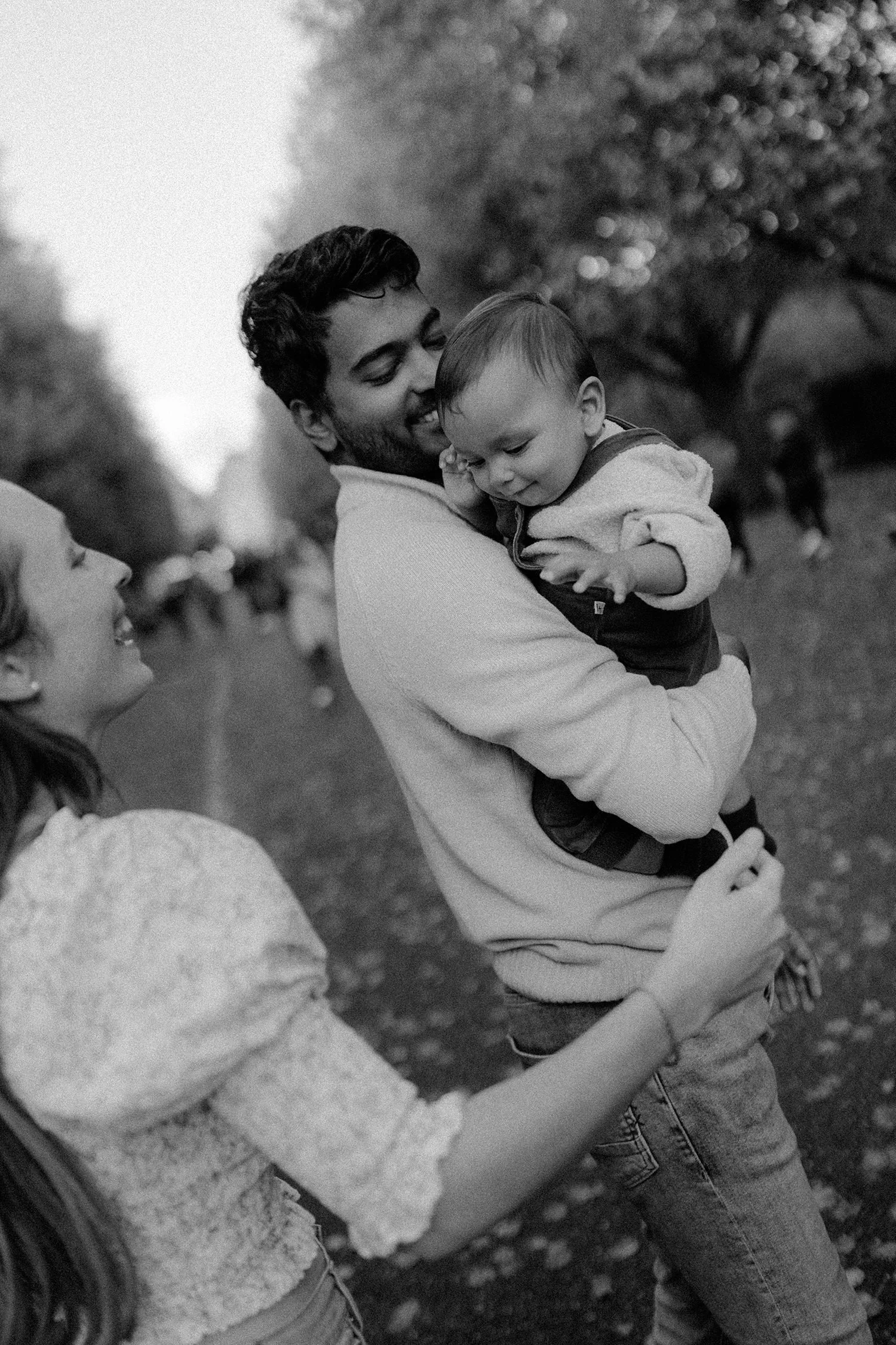 A man with dark hair and a beard holding a young boy in his arms, outdoors with blurred trees in the background. The boy is looking down and the man is smiling with closed eyes, touching his forehead to the boy's.