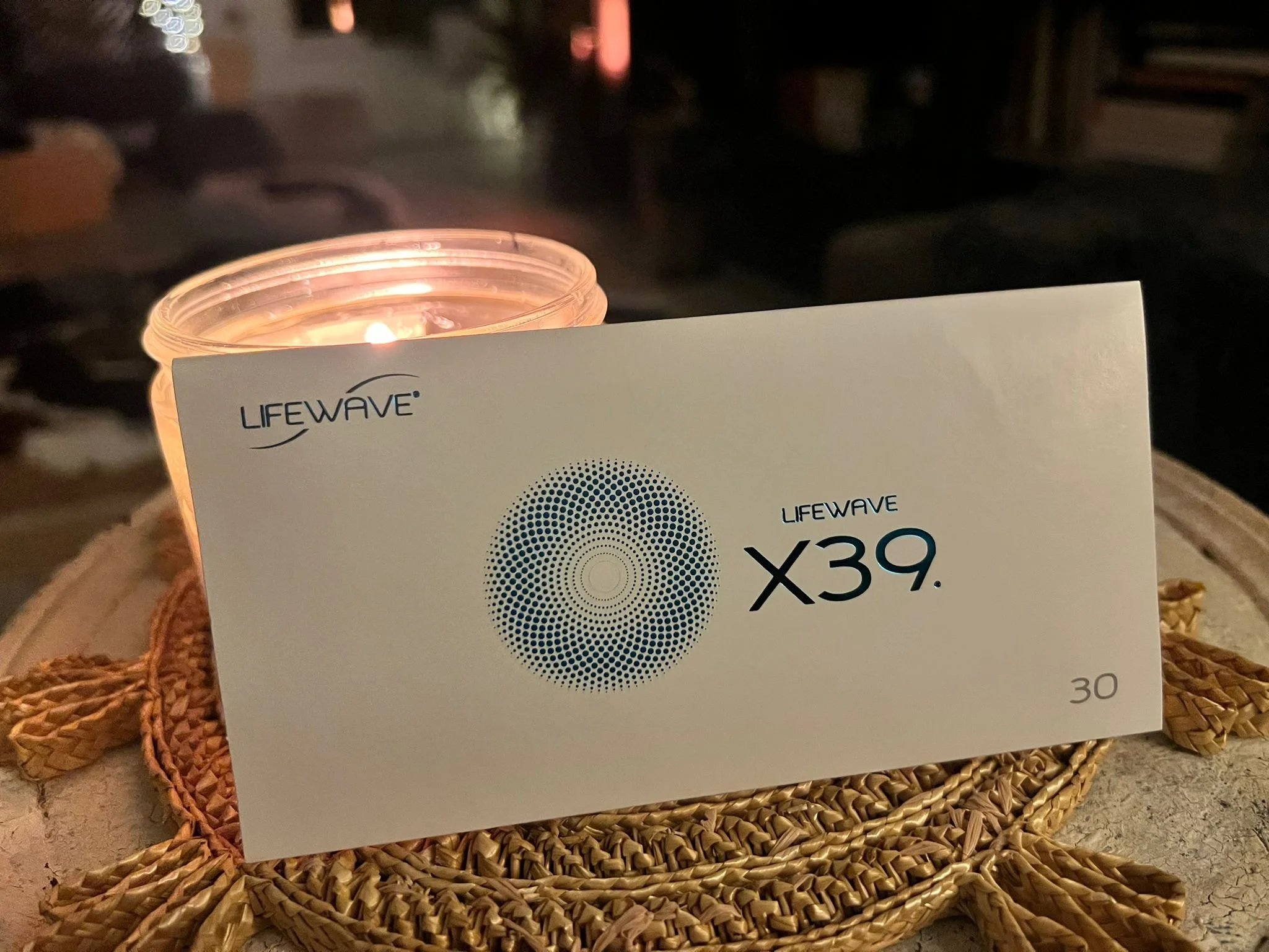 A white LifeWave X39 patch box with a blue circular dot design sits on a woven surface, softly illuminated by a lit candle in a glass jar behind it, creating a warm and calming atmosphere.