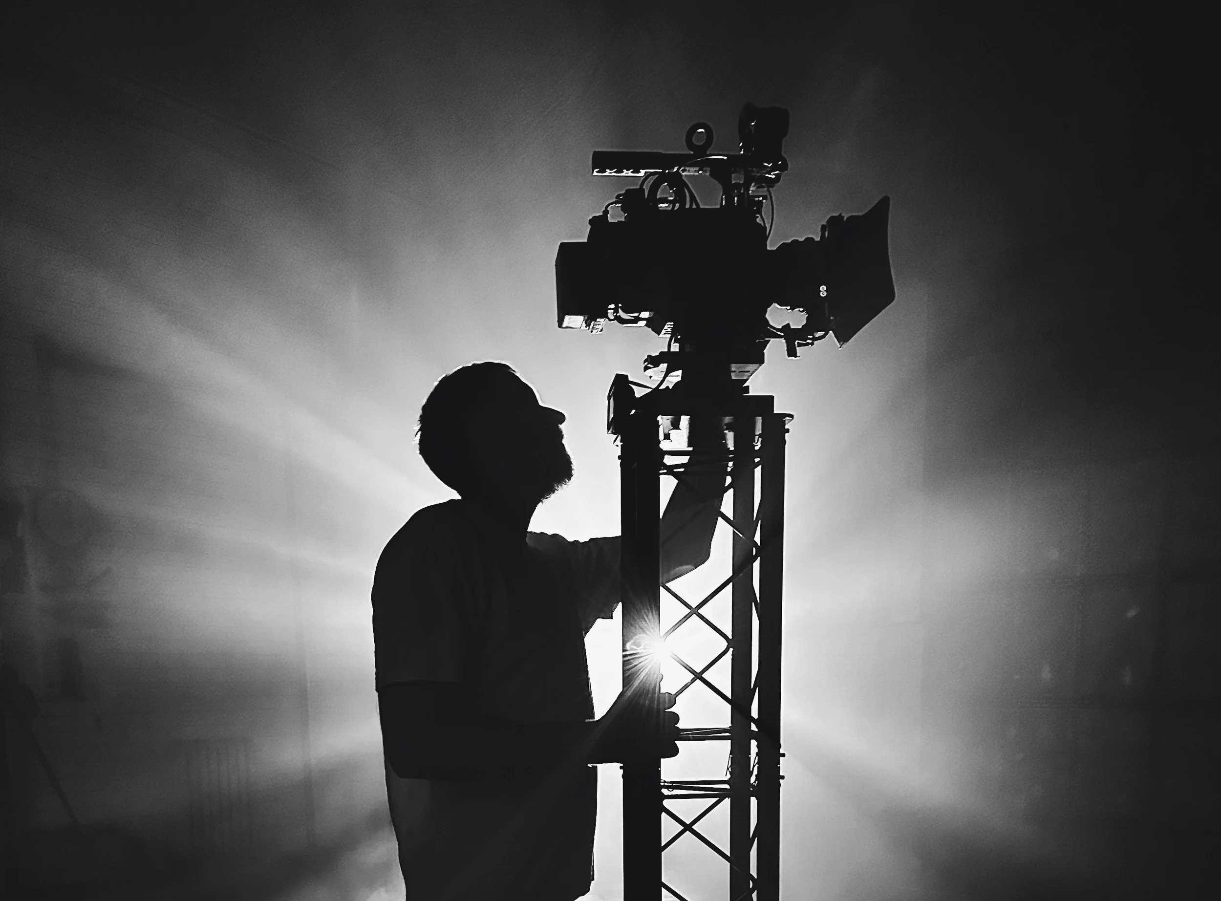 Silhouette of a person with glasses and a beard adjusting equipment on a tall metal stand, with light rays radiating in the background in a black and white setting.