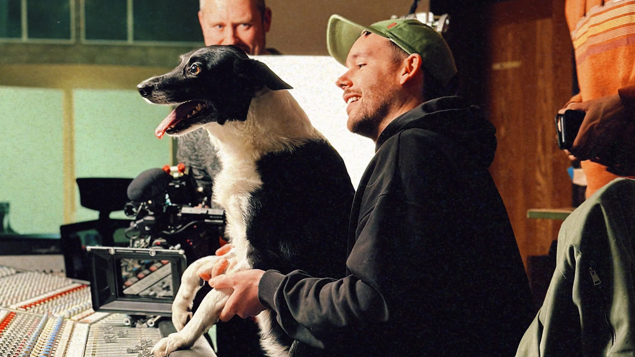 A man holding a black and white dog during a recording session in a studio, with another person in the background near audio equipment.