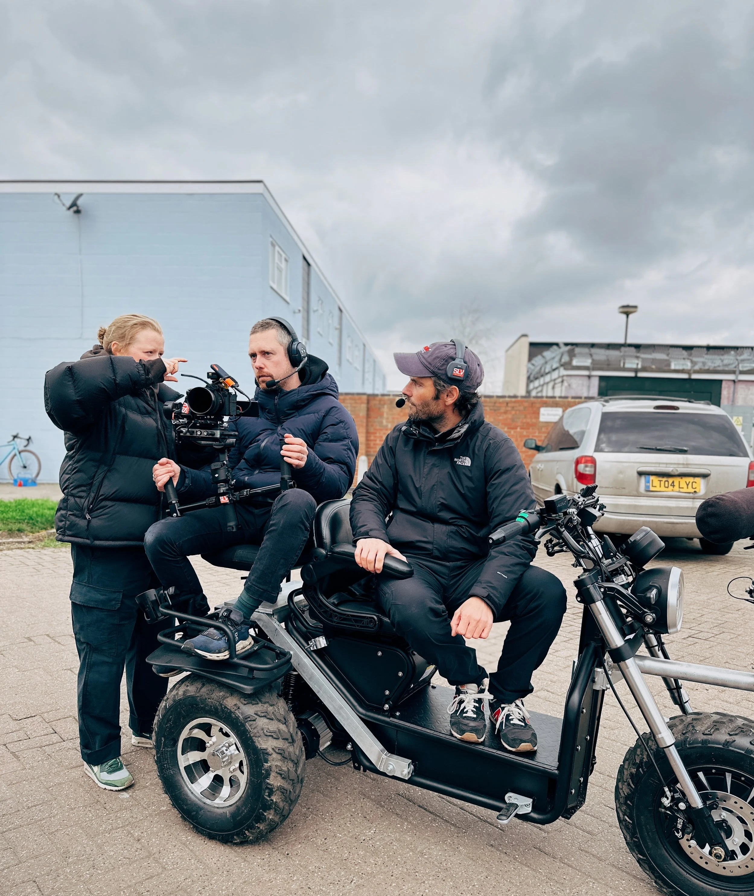 Three people, including a man sitting on an electric scooter and two crew members, preparing for a film shoot outdoors on a cloudy day.