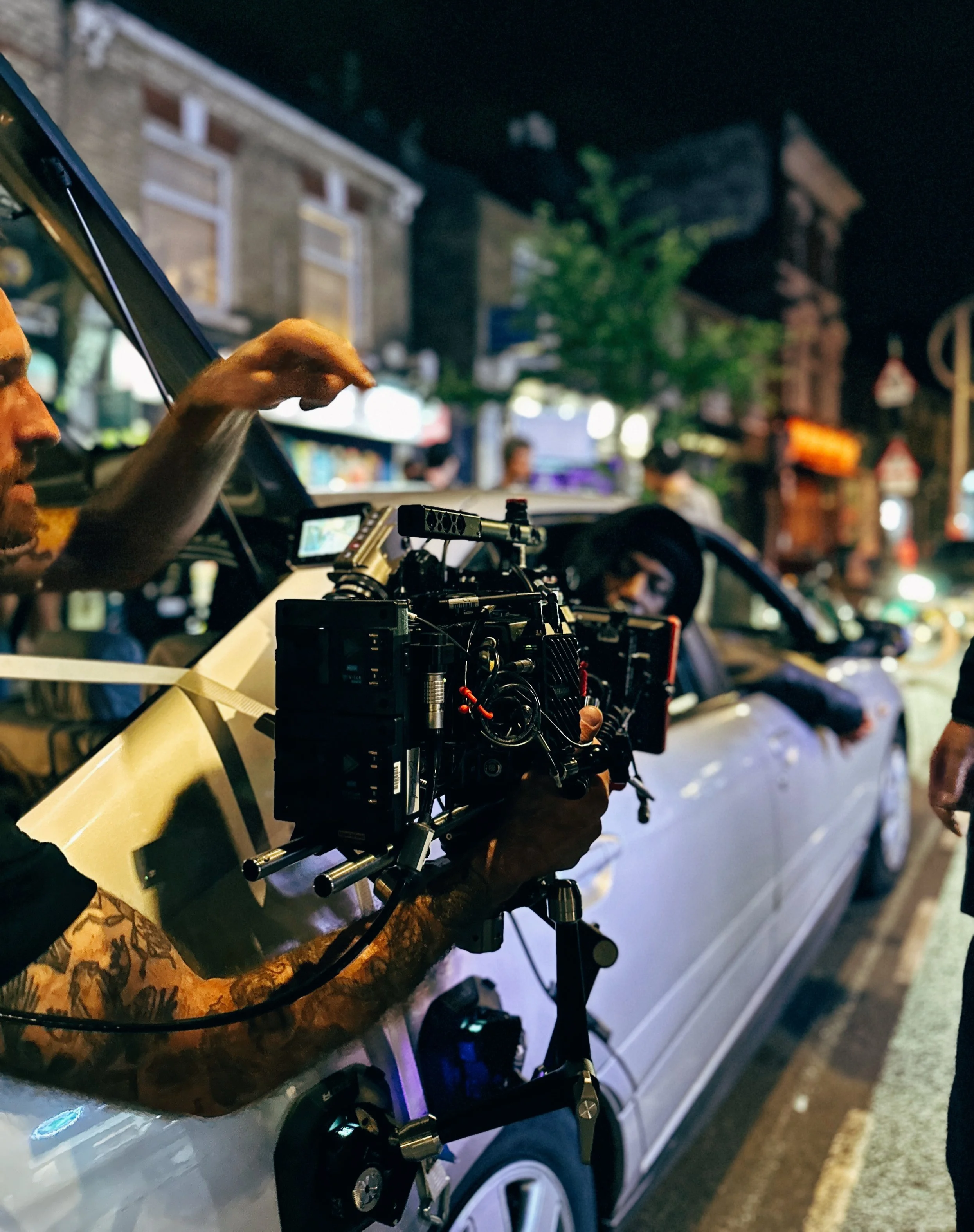 Film crew operating camera equipment inside a white vehicle at night on a city street.