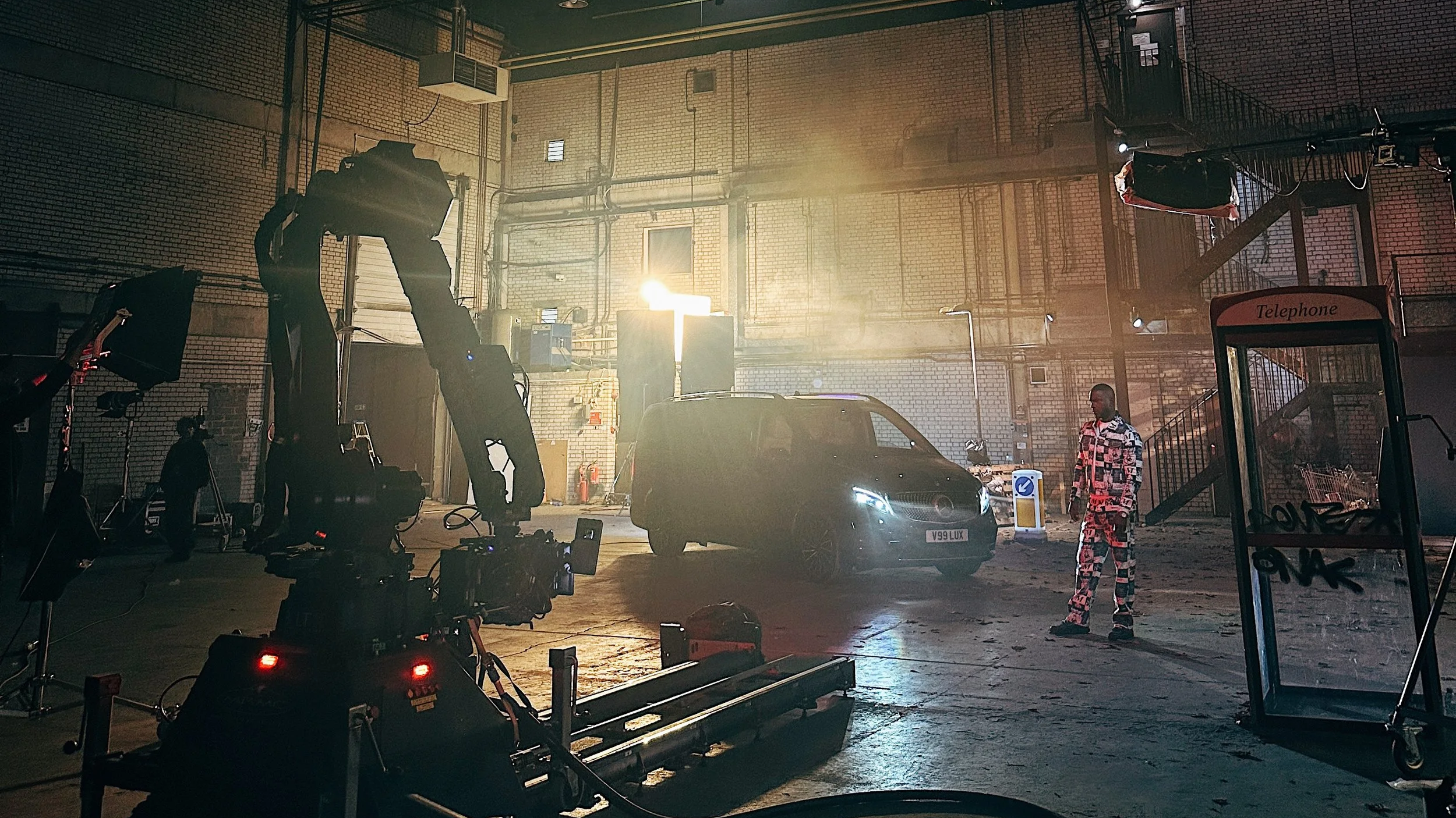 Behind-the-scenes of a film or video shoot in an industrial warehouse, with lighting equipment, a camera crane, a person in patterned clothes near a black car, and a telephone booth.
