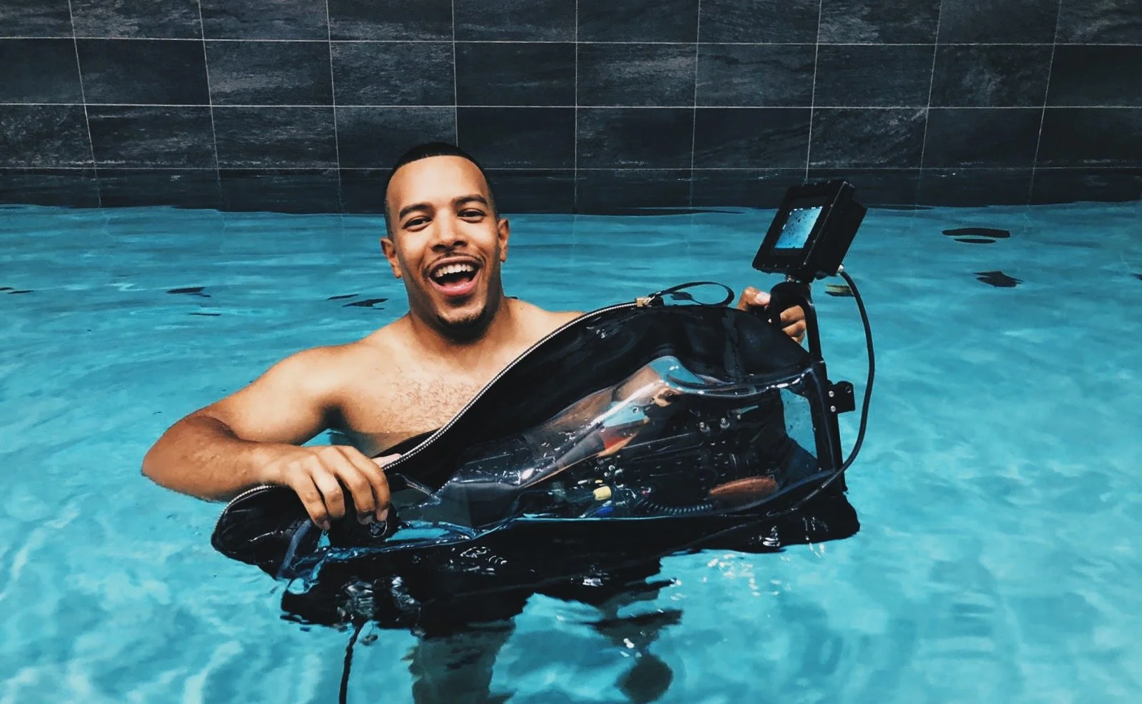 A smiling man in a swimming pool holding underwater photography equipment.