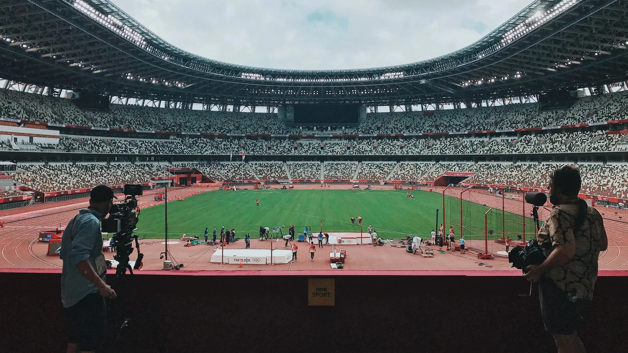 View of a large athletic stadium with ongoing track and field event. Two cameramen are seen in the foreground, one on the left and one on the right, capturing the scene. The stadium has a seating area with tiered rows and a green field in the center,