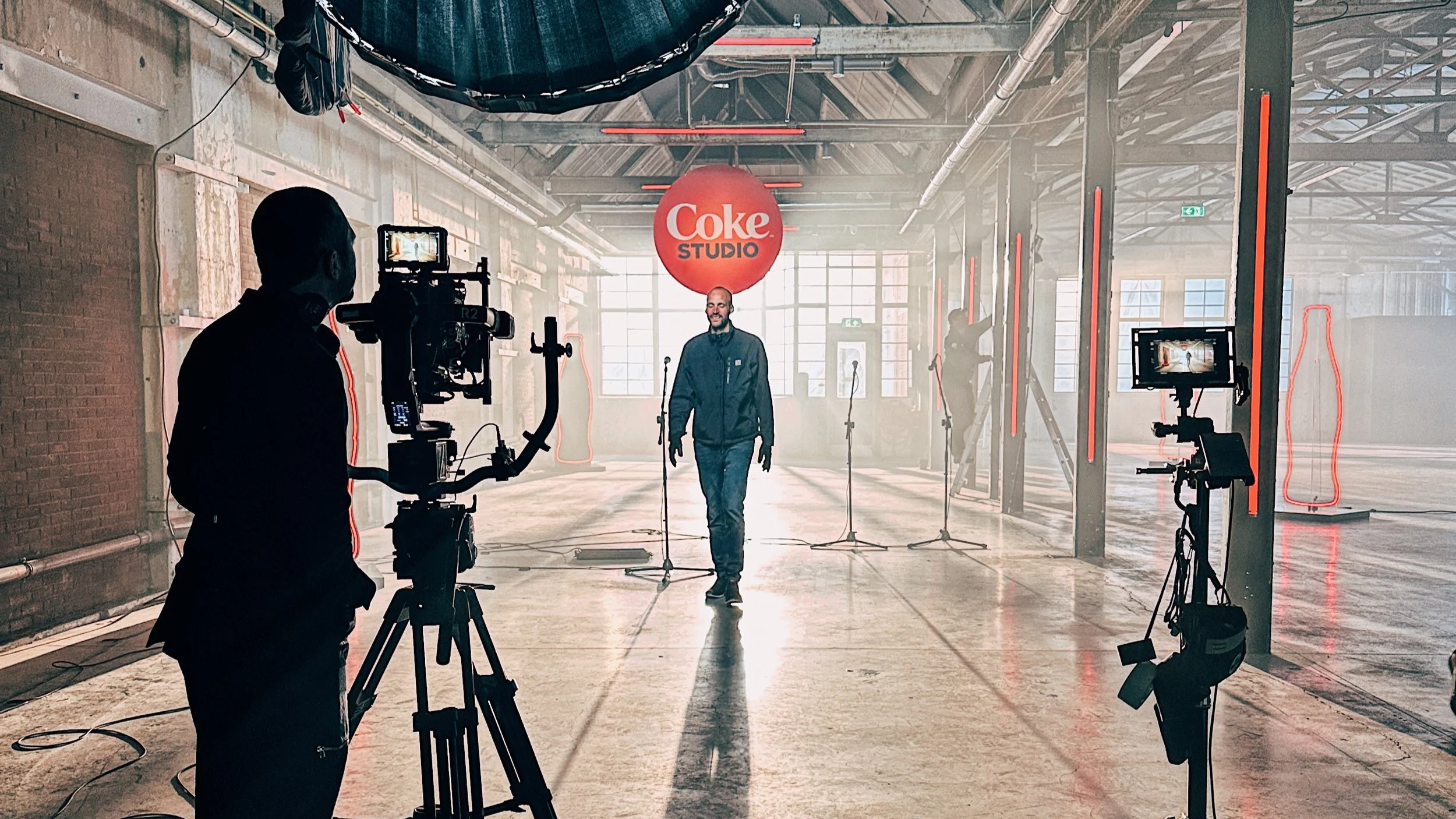 A man walking in a spacious, industrial-style studio with a Coca-Cola sign reading 'Coke Studio' hanging from the ceiling. Cameras and crew members are filming him, with bright natural light coming through large windows in the background.