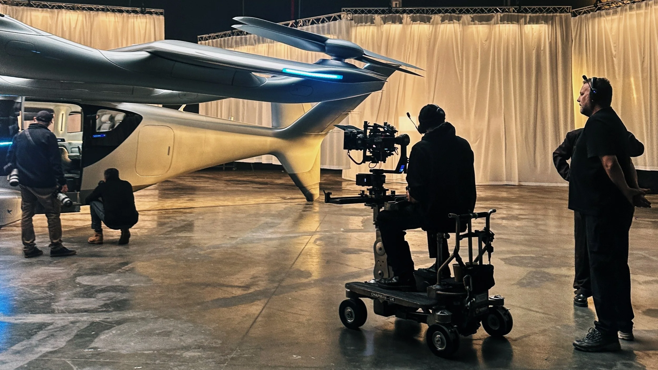 Filming crew working on a spaceship set indoors, with two people near the spaceship and a person operating a camera.
