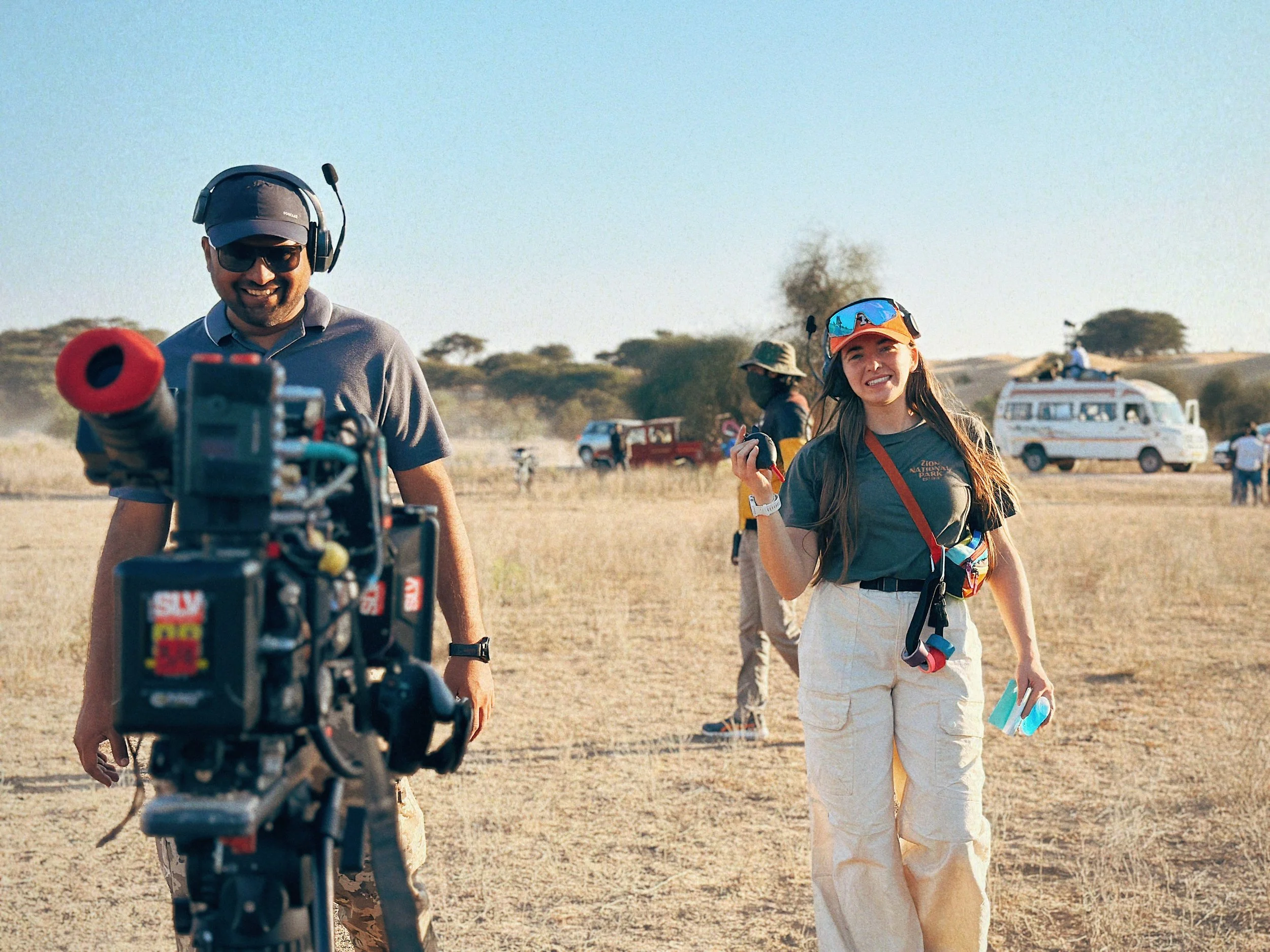 Two people, a man and a woman, are standing in a dry, open field on a sunny day. The man is smiling and wearing sunglasses, headphones, a gray polo shirt, and camouflage pants, standing behind a camera setup. The woman is smiling, holding a camera an