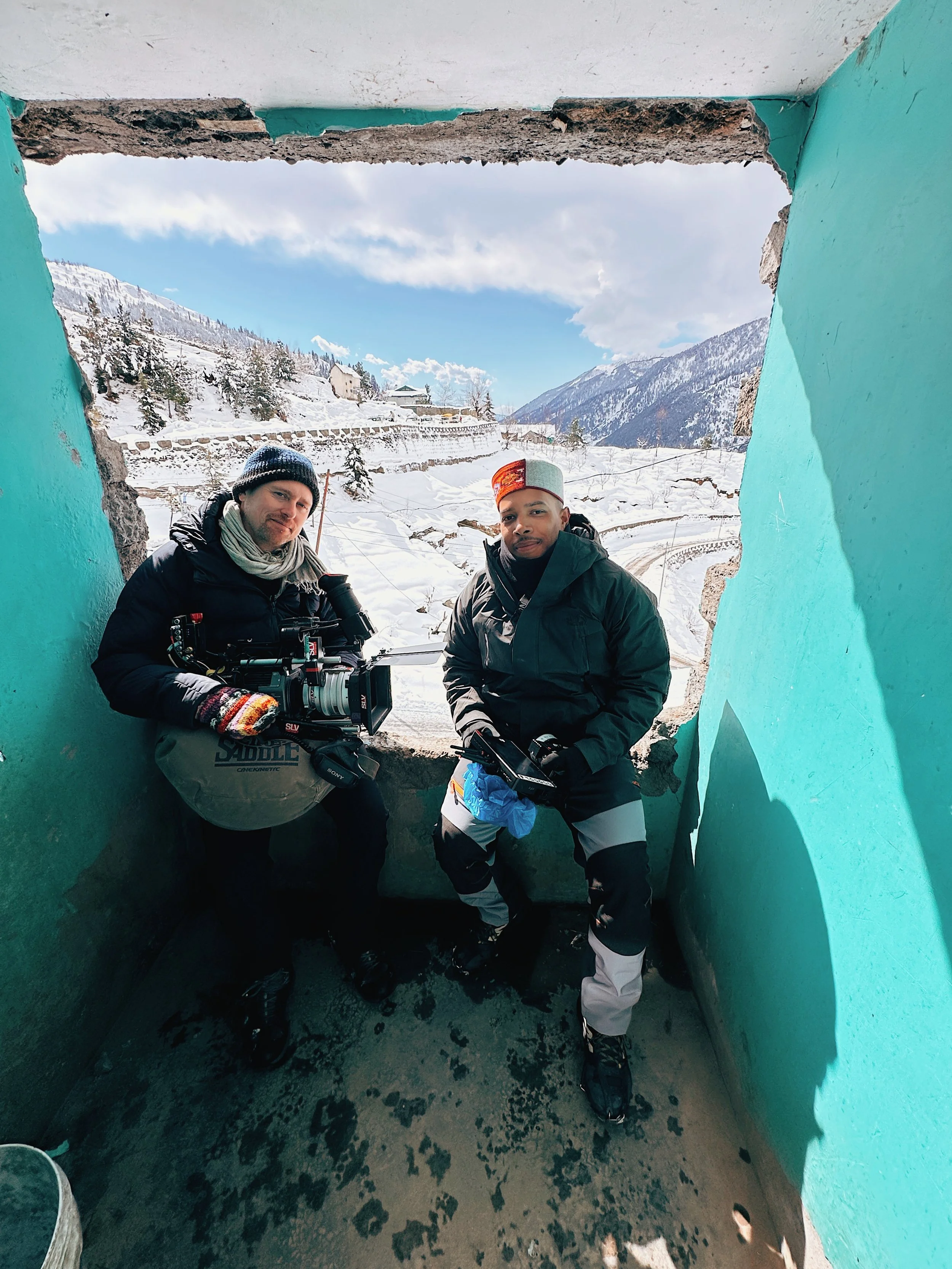 Two men filming with a camera in a partially destroyed structure with snow-covered mountains and houses in the background.