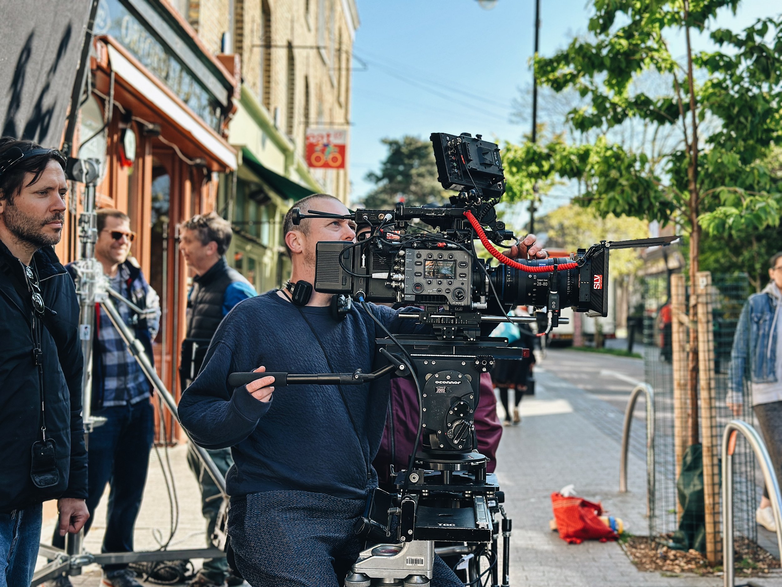 A film crew is filming on a busy city street. A camera operator is adjusting a professional film camera mounted on a tripod, with a director or crew member standing nearby. In the background, several people are walking along the sidewalk, with some s