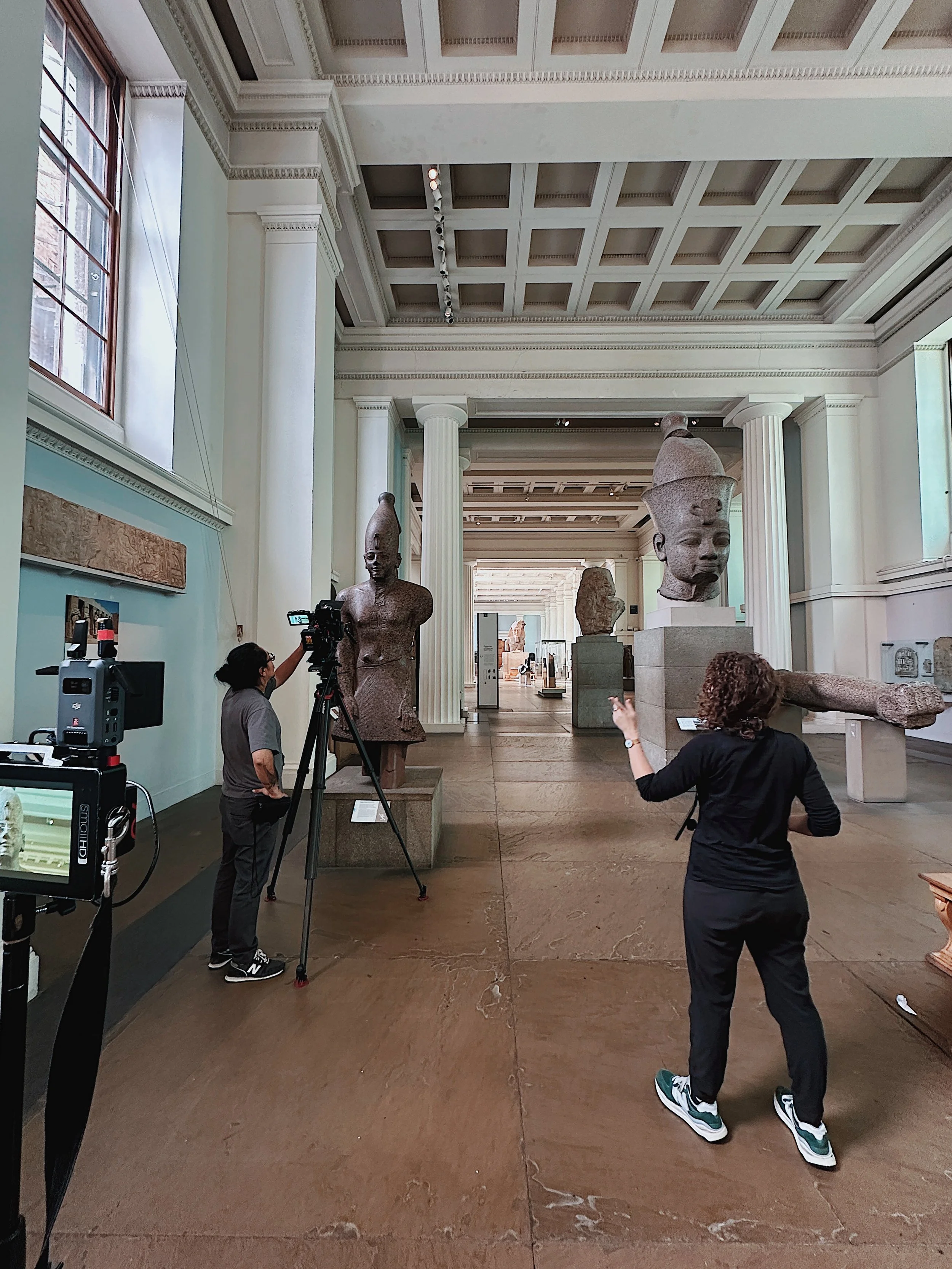 People in a museum viewing ancient Egyptian sculptures, including a large head of a pharaoh and a partially missing statue of a figure with a headdress.