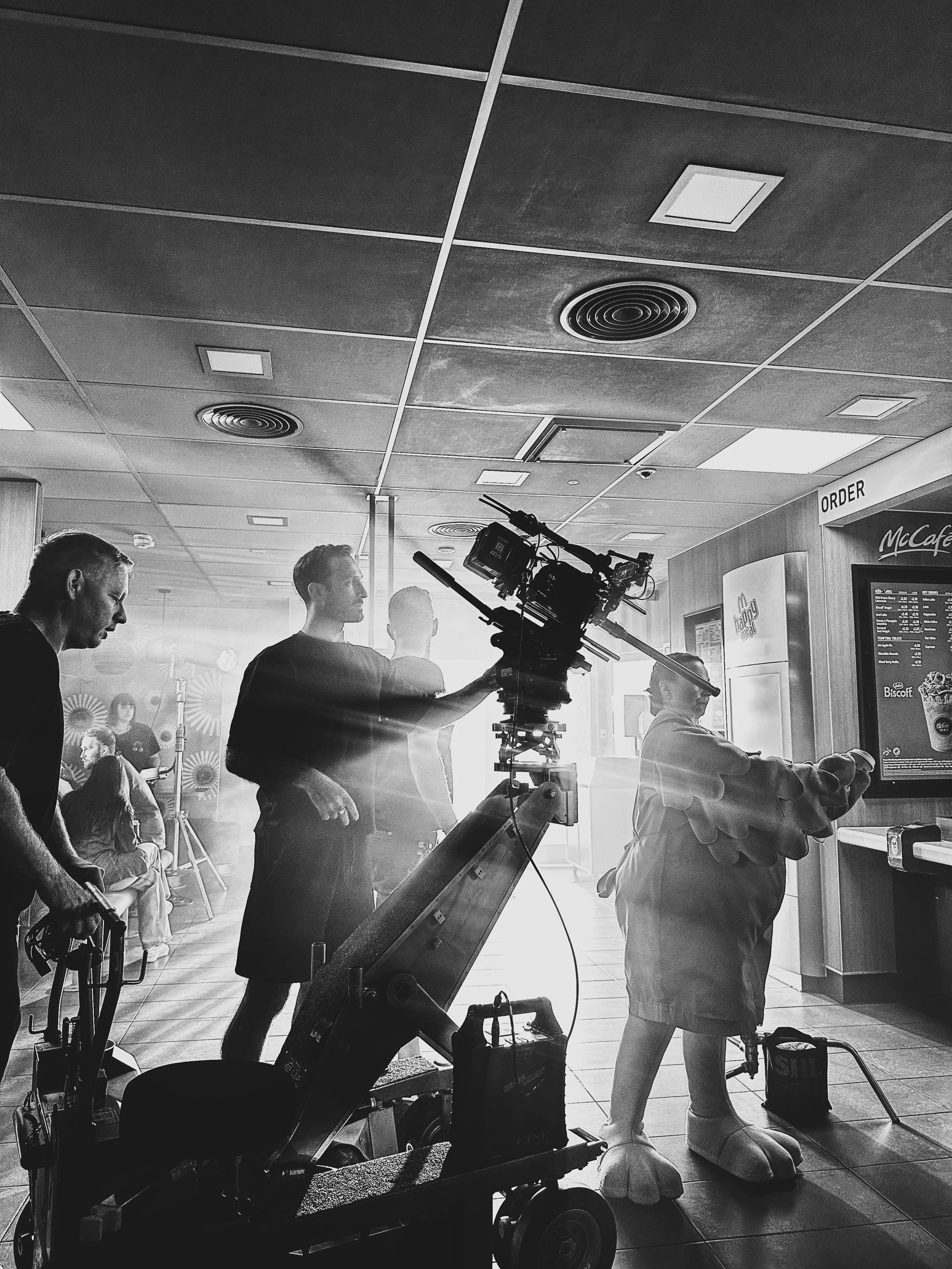 Filming of a commercial or scene at a fast food restaurant, with a camera on a dolly and crew members, one of whom is wearing a costume resembling a chicken or mascot, reaching for the menu.