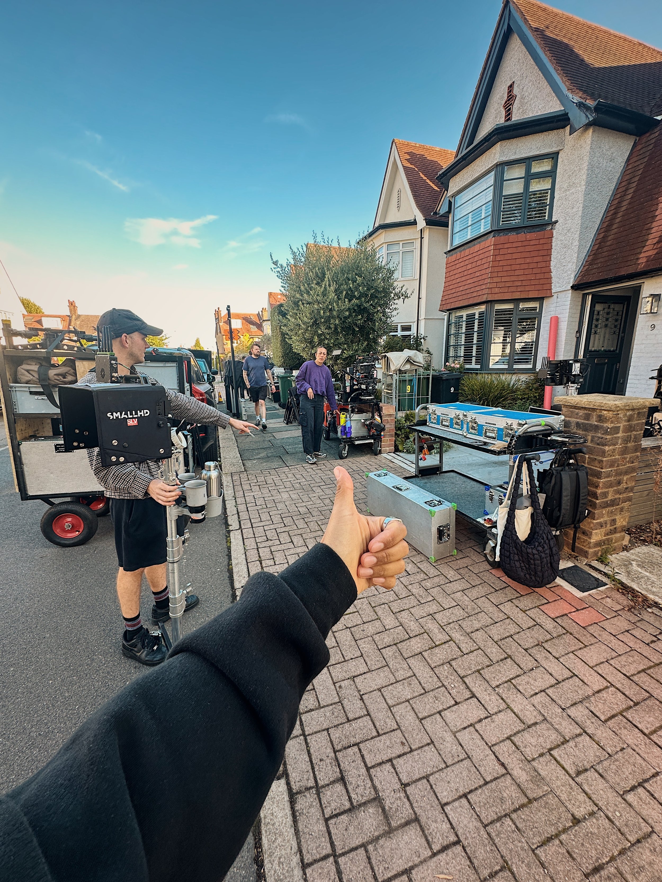 Filming crew setting up equipment outside a house on a suburban street, with a person giving a thumbs-up in the foreground.