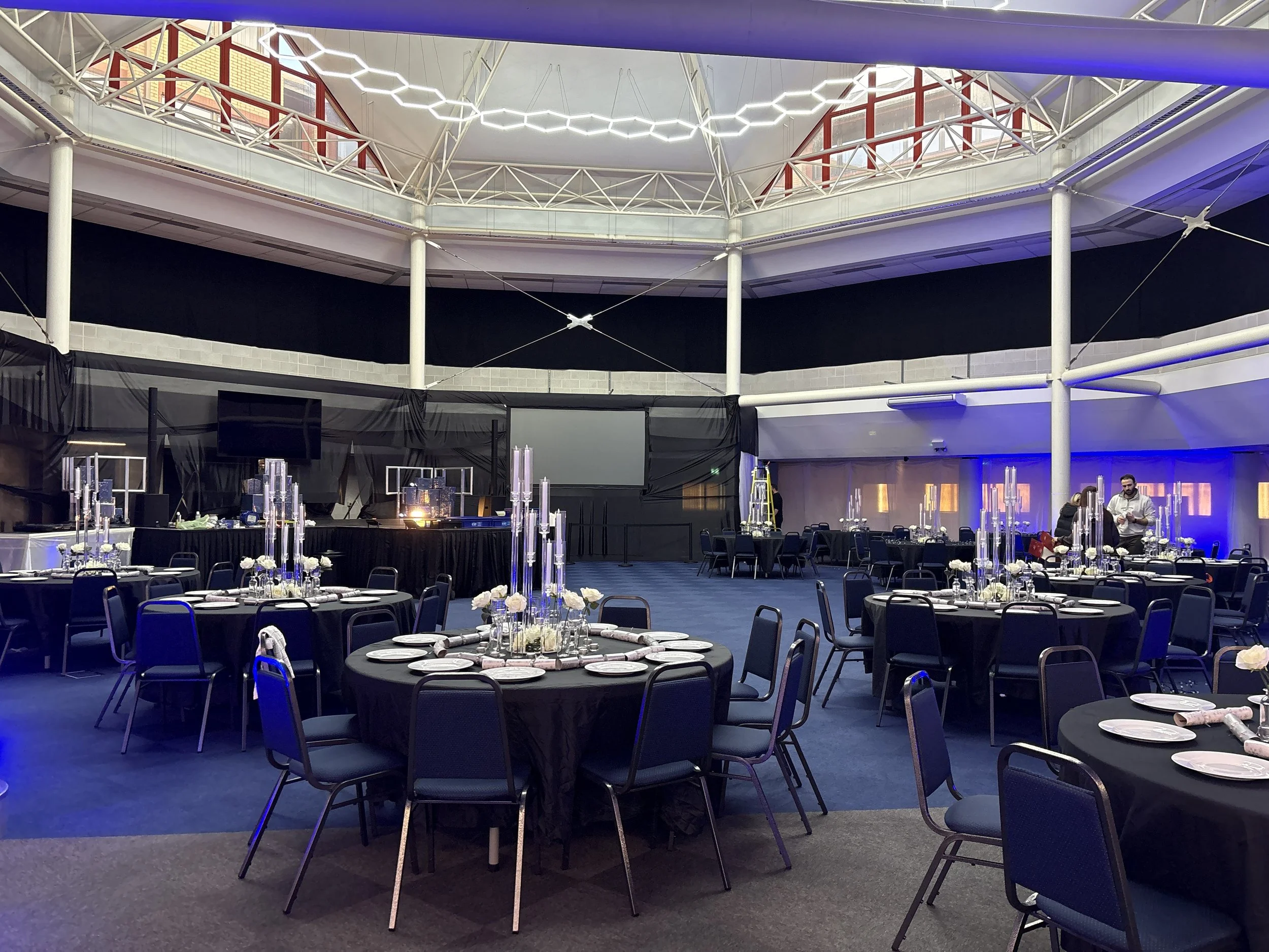 Banquet hall with round tables covered in black tablecloths, decorated with tall candle holders and flower arrangements, set for a formal event, with a stage and large screens at the front and some staff preparing in the background.