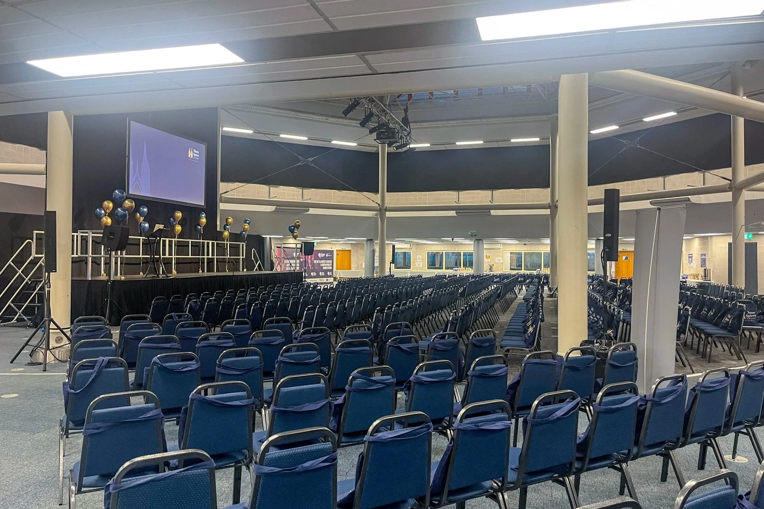A large indoor conference or event space set up with rows of blue chairs facing a stage with a large screen, balloons, and speakers.