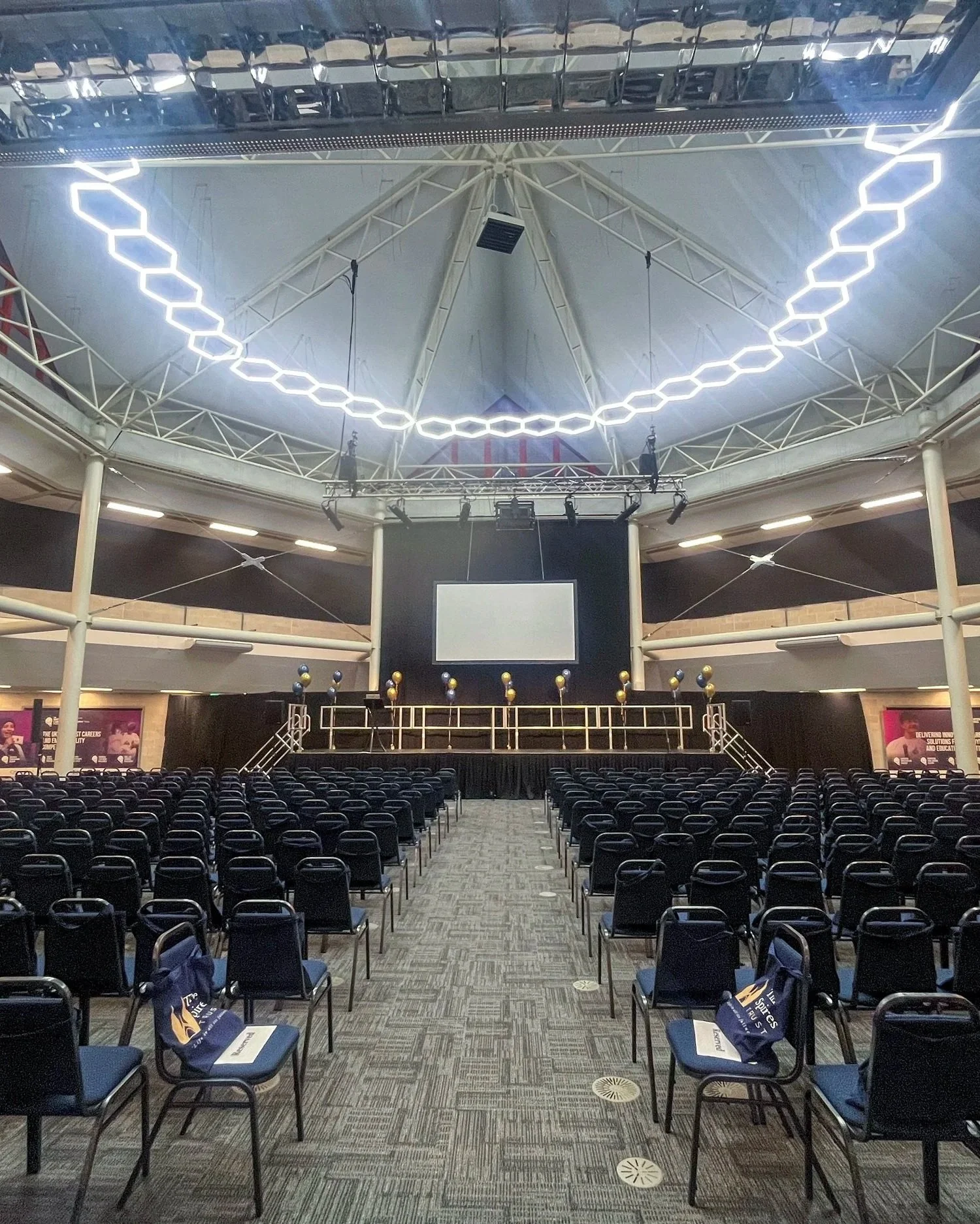 Large conference hall with rows of black chairs facing a stage with a large screen and a black backdrop, decorated with yellow and black balloons, under a high ceiling with modern light fixtures.