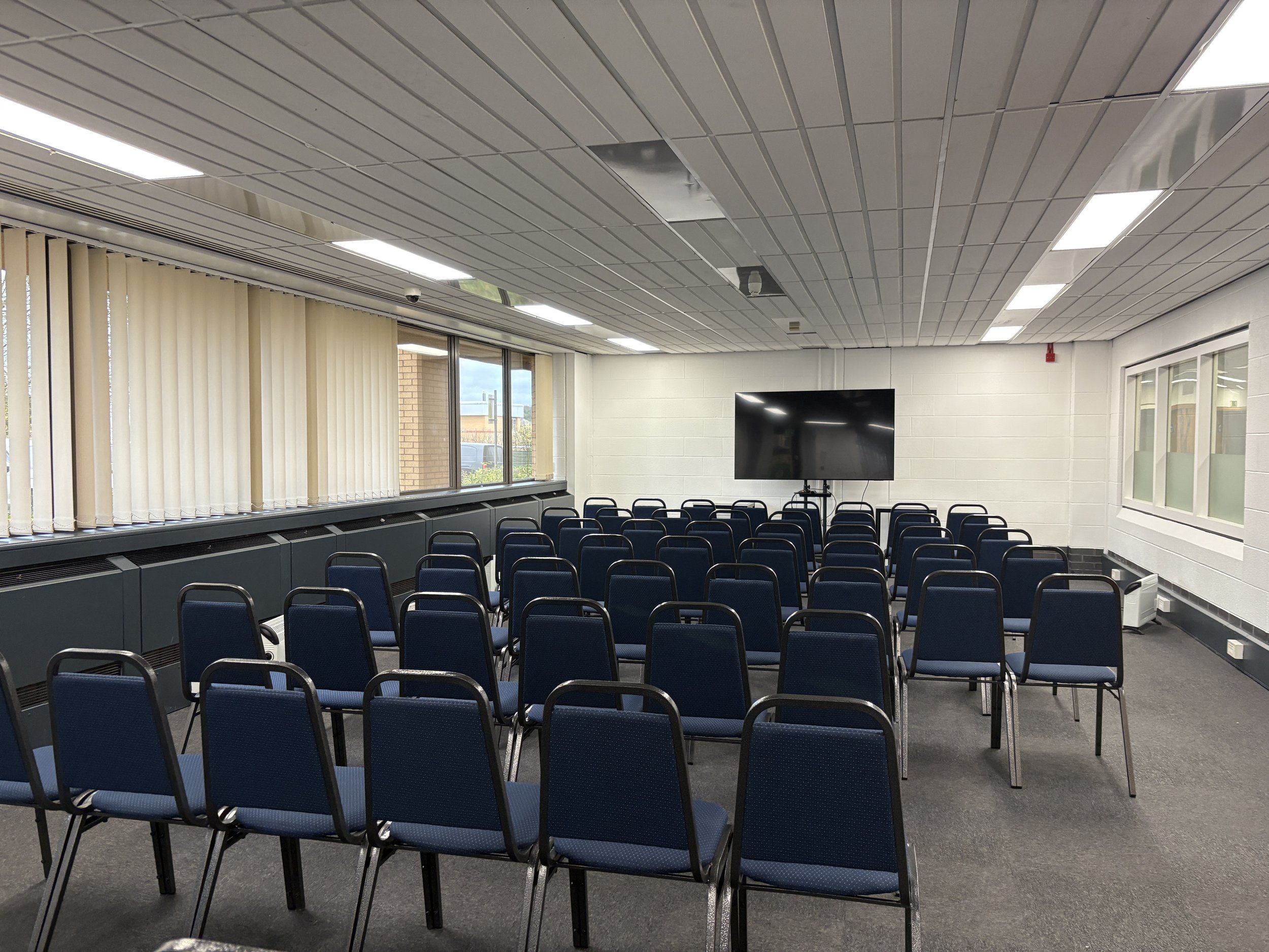 Empty classroom with rows of blue chairs facing a large flat screen TV, white brick walls, and windows with vertical blinds.