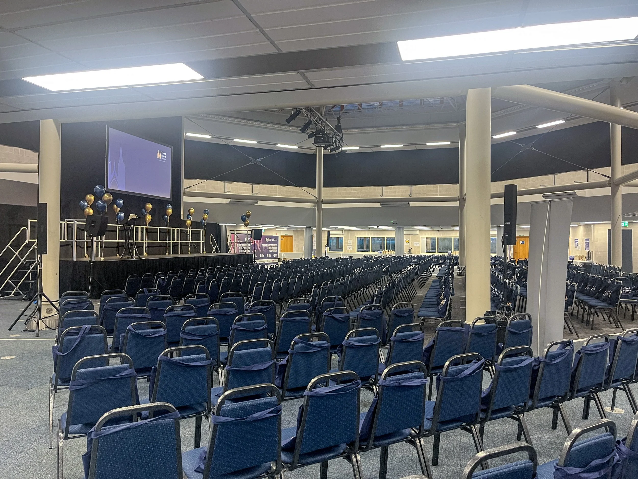 Empty conference hall with rows of blue chairs, a stage with decorations, and a large screen, prepared for an event.