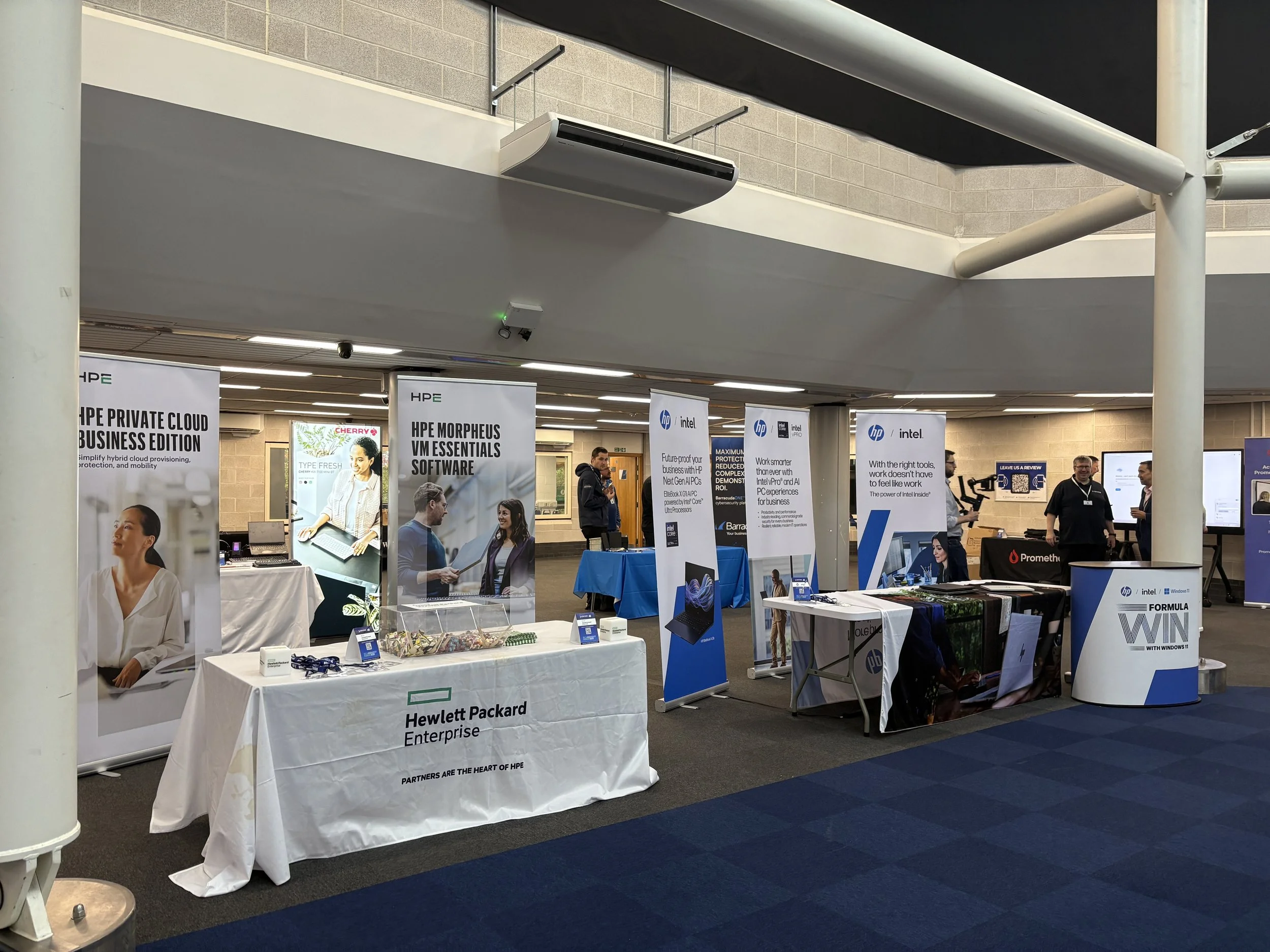 Technology booths set up at an indoor convention or trade show with banners, tables, and promotional materials for different tech companies like Hewlett Packard Enterprise, HP, and Intel.