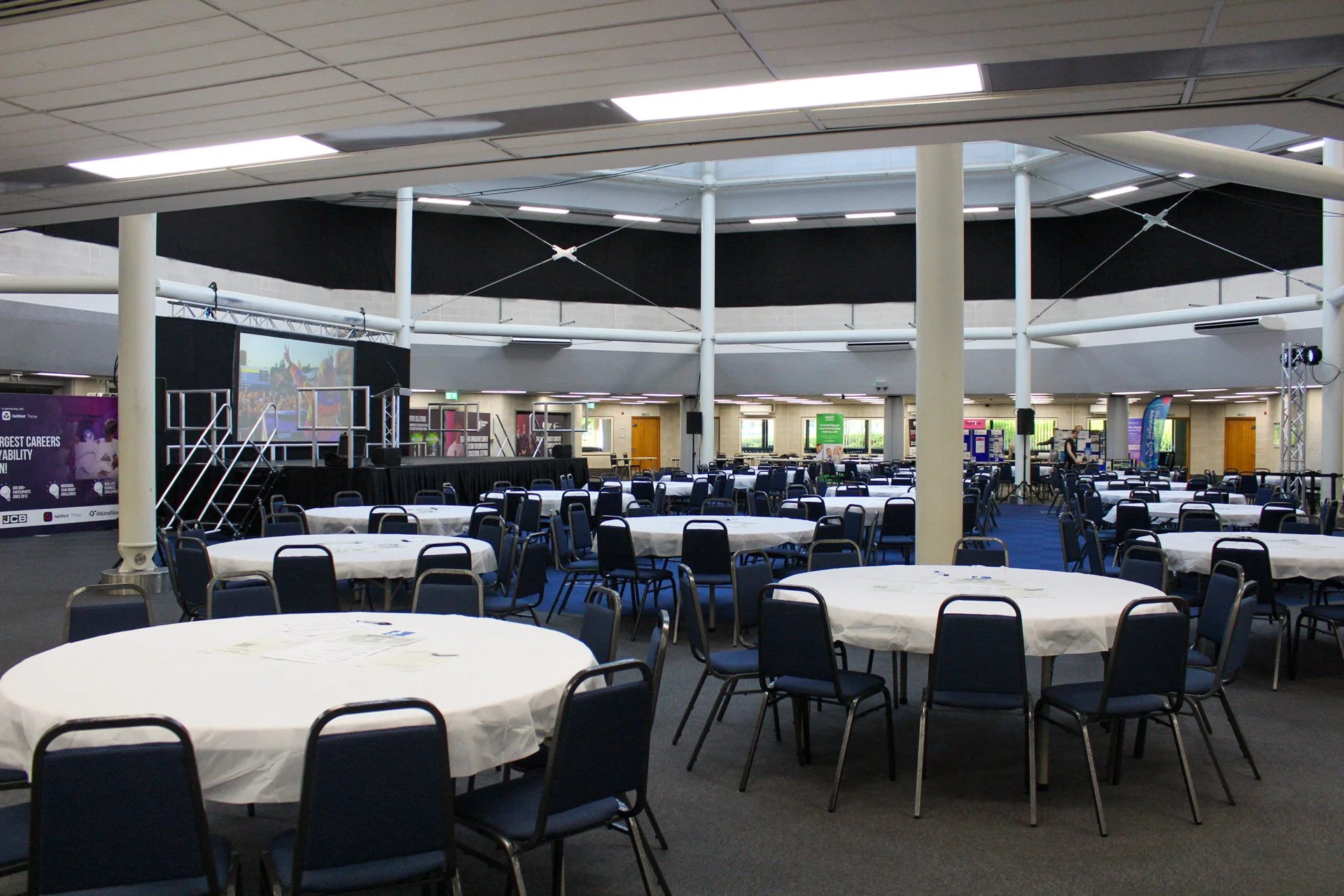 Round tables with white tablecloths and blue chairs set up in a large event hall with a stage, screens, and banners.