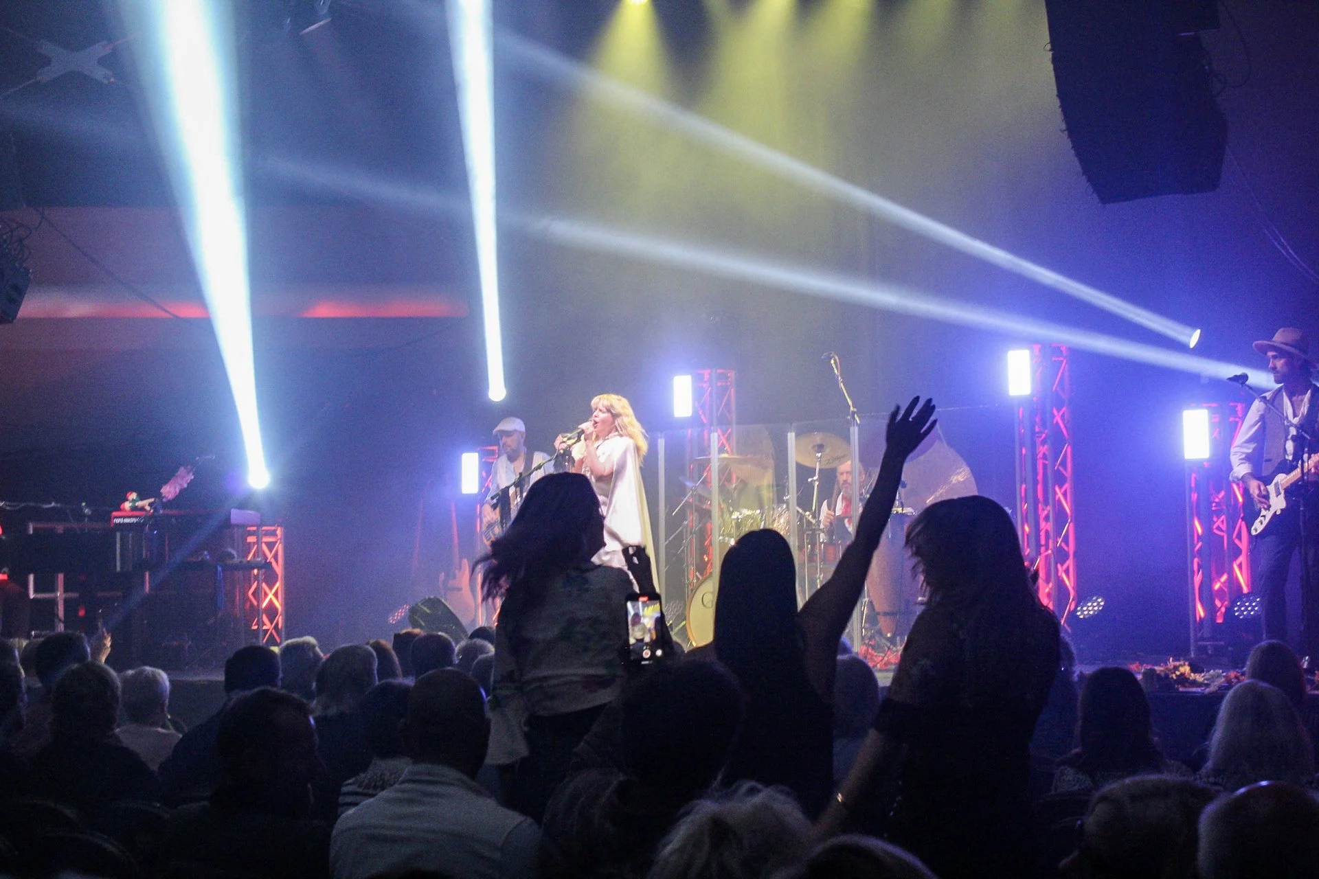 Live concert with a band performing on stage, colorful lights and audience members, including a woman raising her hand.
