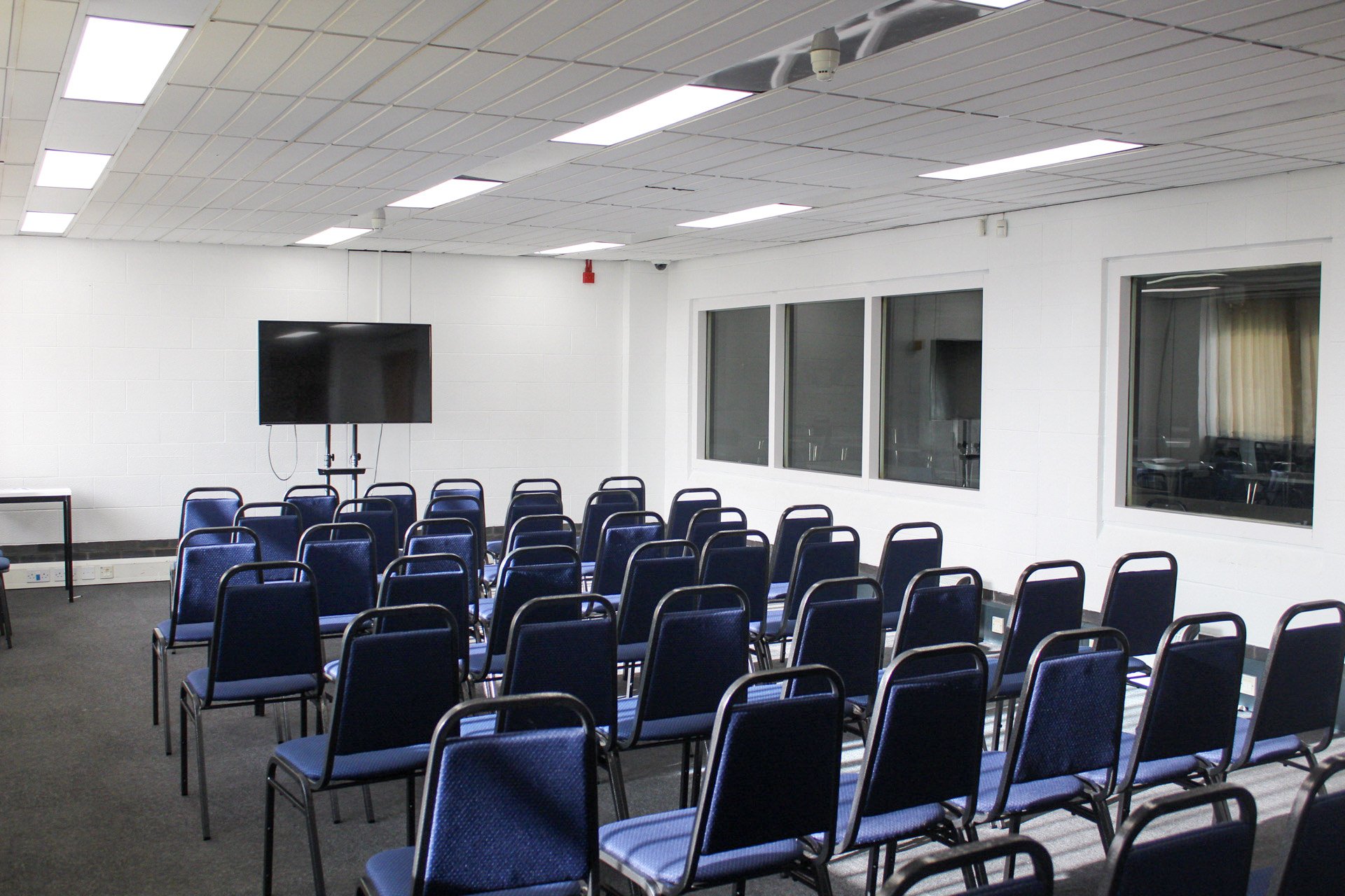 Empty conference room with multiple rows of blue chairs, a large wall-mounted television, white cinder block walls, and windows on one wall.