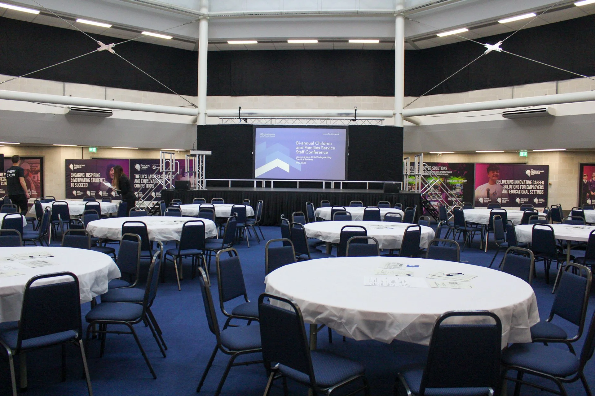 Empty conference room with round tables covered with white tablecloths, blue chairs, a stage with a large screen displaying a presentation slide, and posters on the walls.