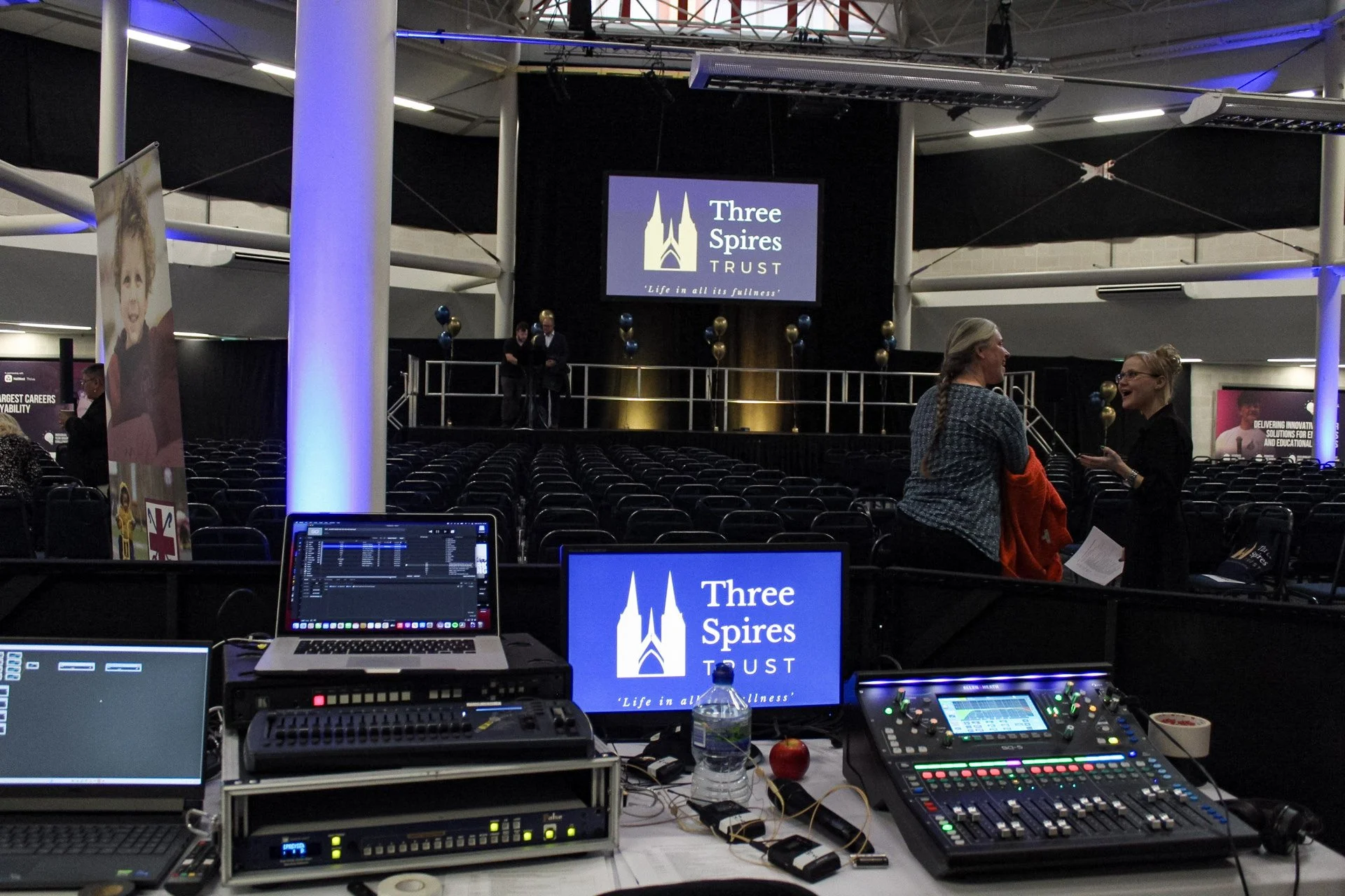 Event stage with large screens displaying 'Three Spires TRUST' logo and slogan in an auditorium. In front, audio and lighting control equipment with laptops, soundboards, and a water bottle, apple, and miscellaneous items. Two women are conversing an