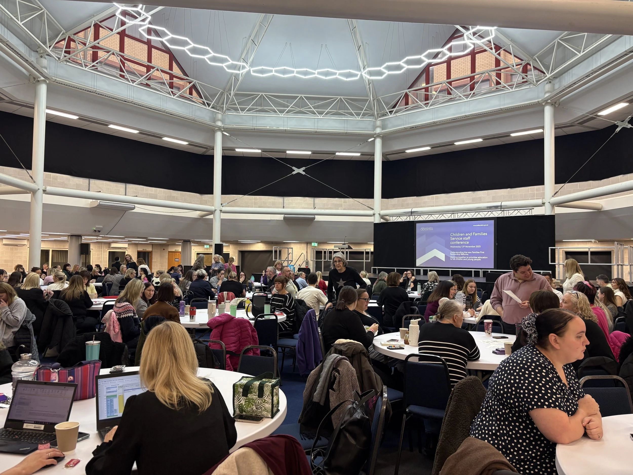 Large conference room filled with many people seated at round tables attending a presentation on children and families, with a large screen displaying the conference information.