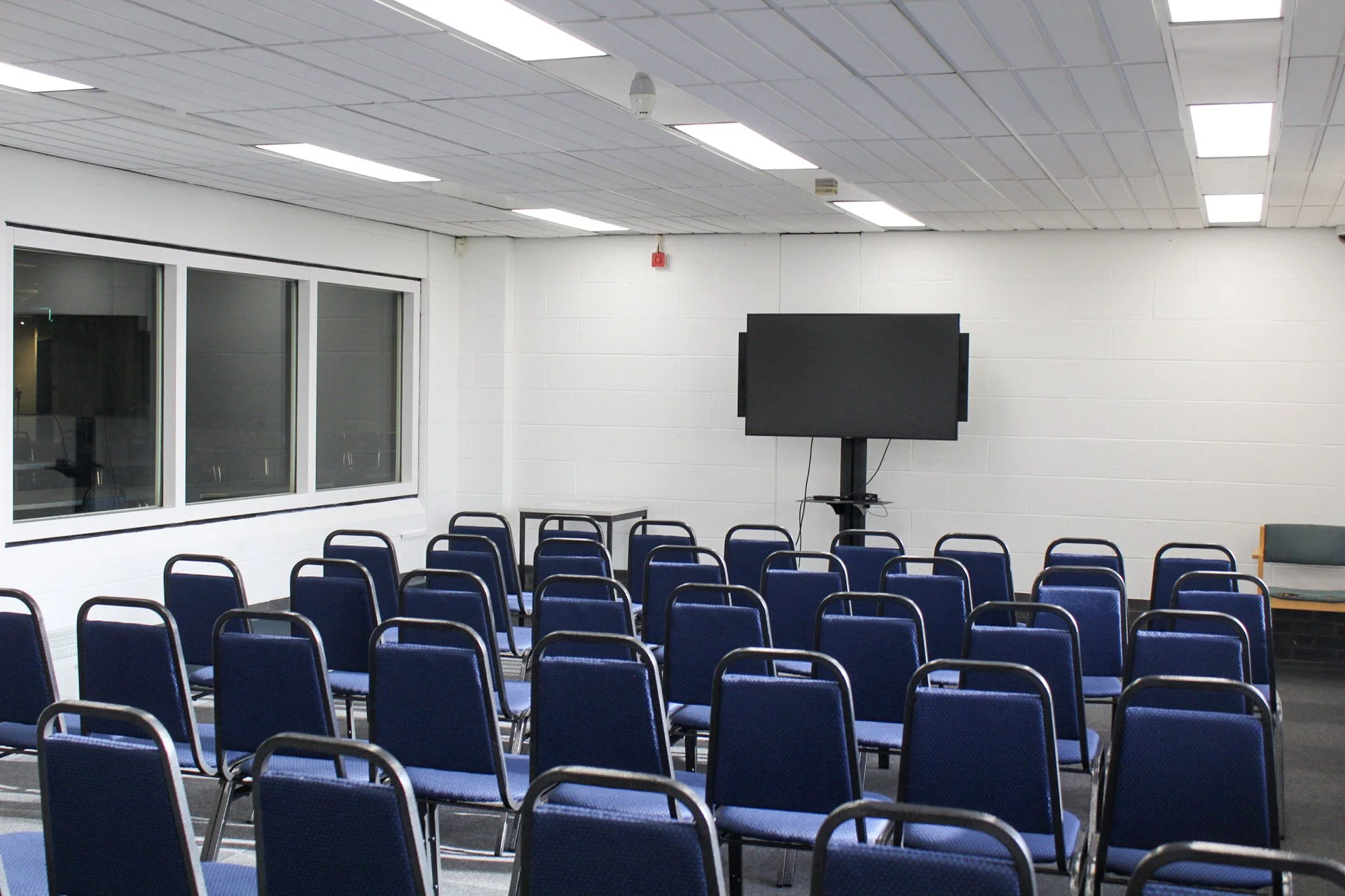 Empty room set up for presentation with rows of blue chairs facing a large black screen on a stand, white walls, windows on the left, and ceiling lights.