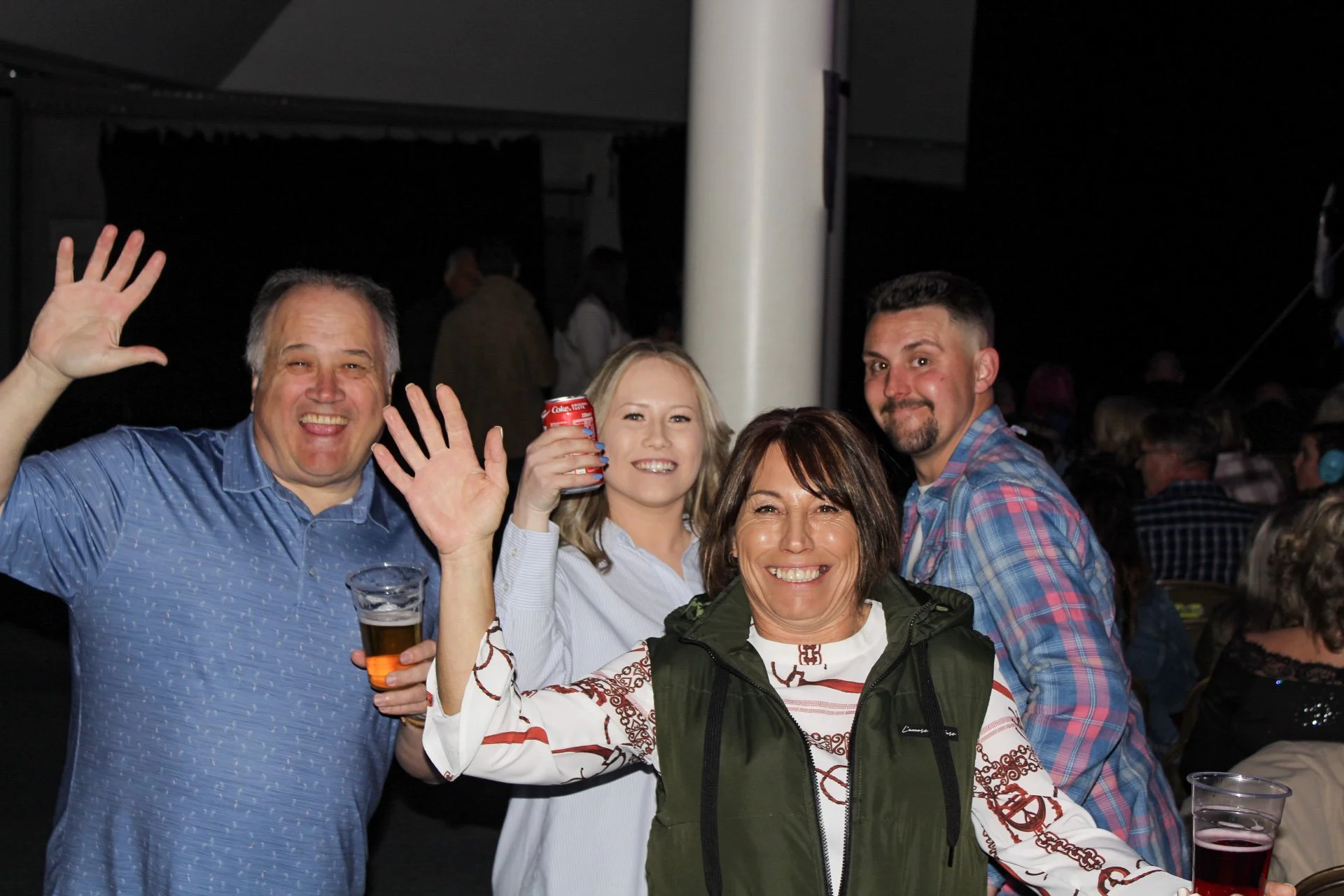 Group of four people smiling and raising their hands at a social gathering, holding drinks, in a dark indoor setting.
