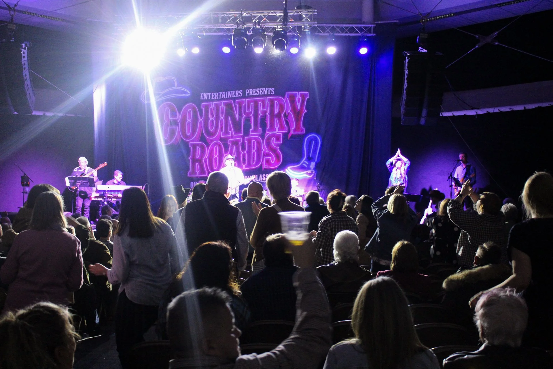 An audience watching a country music concert on a stage with a large banner that reads 'Country Roads' and performing musicians.