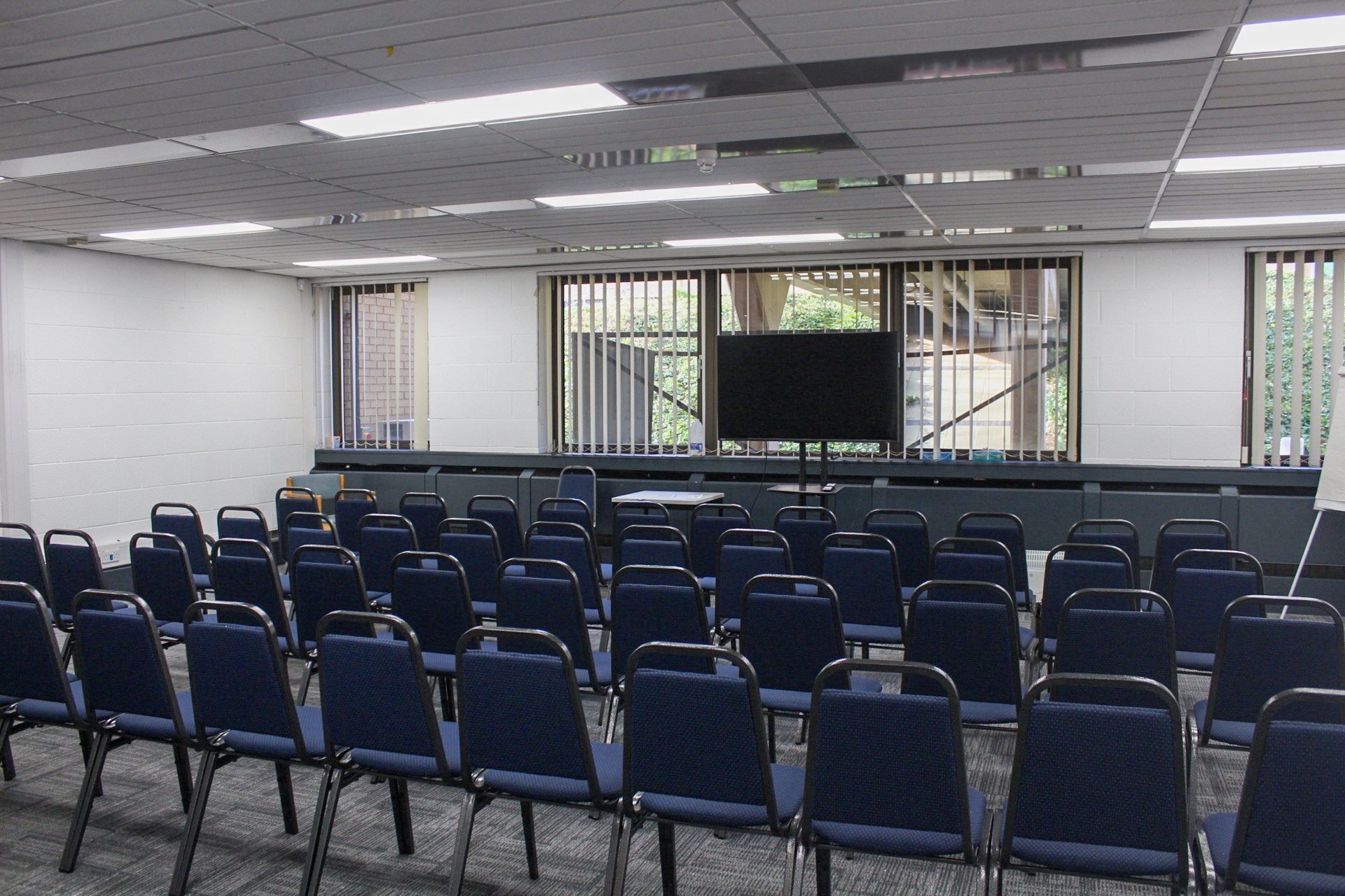 Empty conference room with rows of blue chairs facing a large screen, windows with vertical blinds allowing natural light, and a gray carpeted floor.