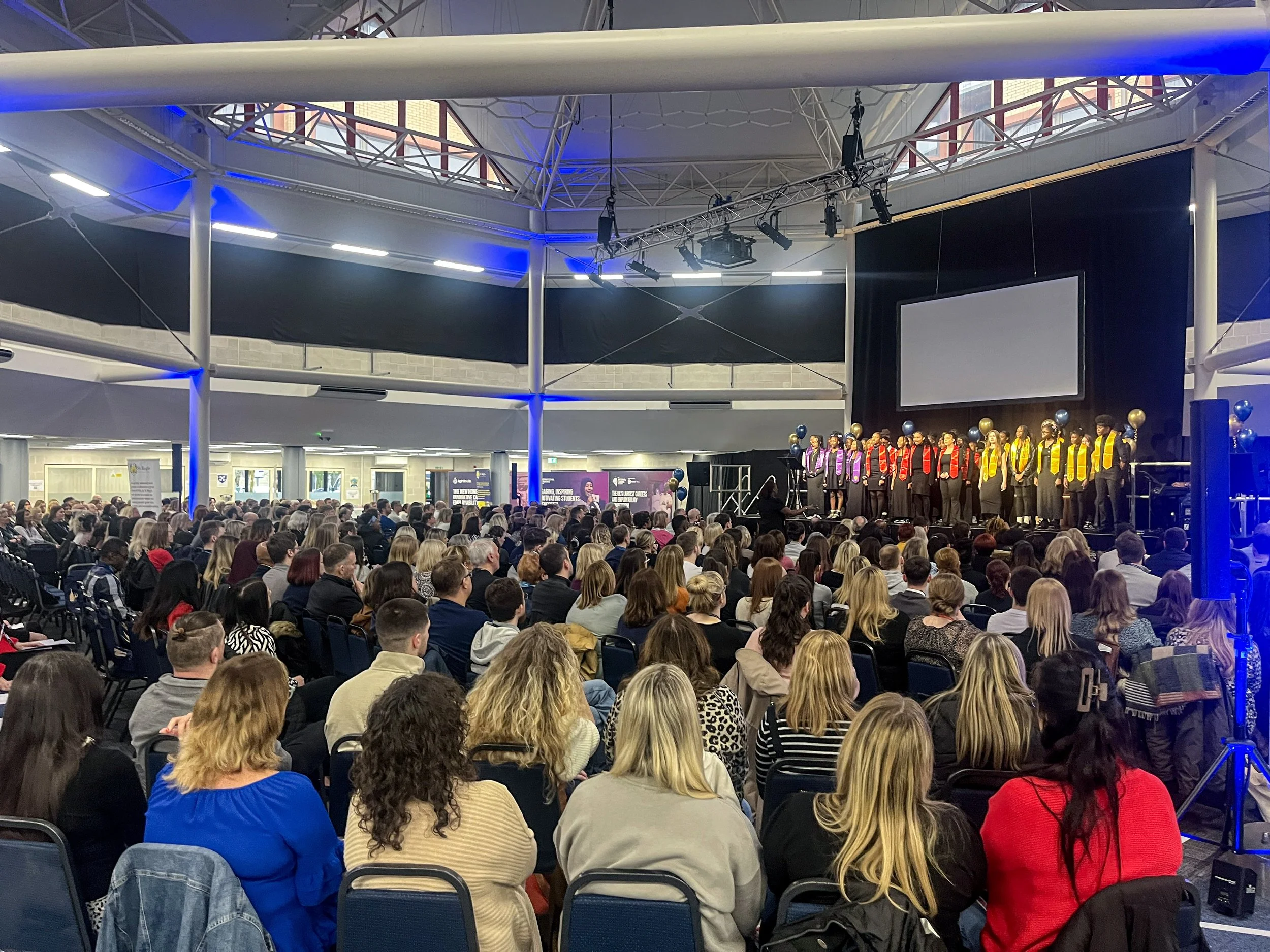 Large audience watching a choir perform on stage in a spacious indoor venue with high ceilings, decorated with balloons and a large screen above the choir.