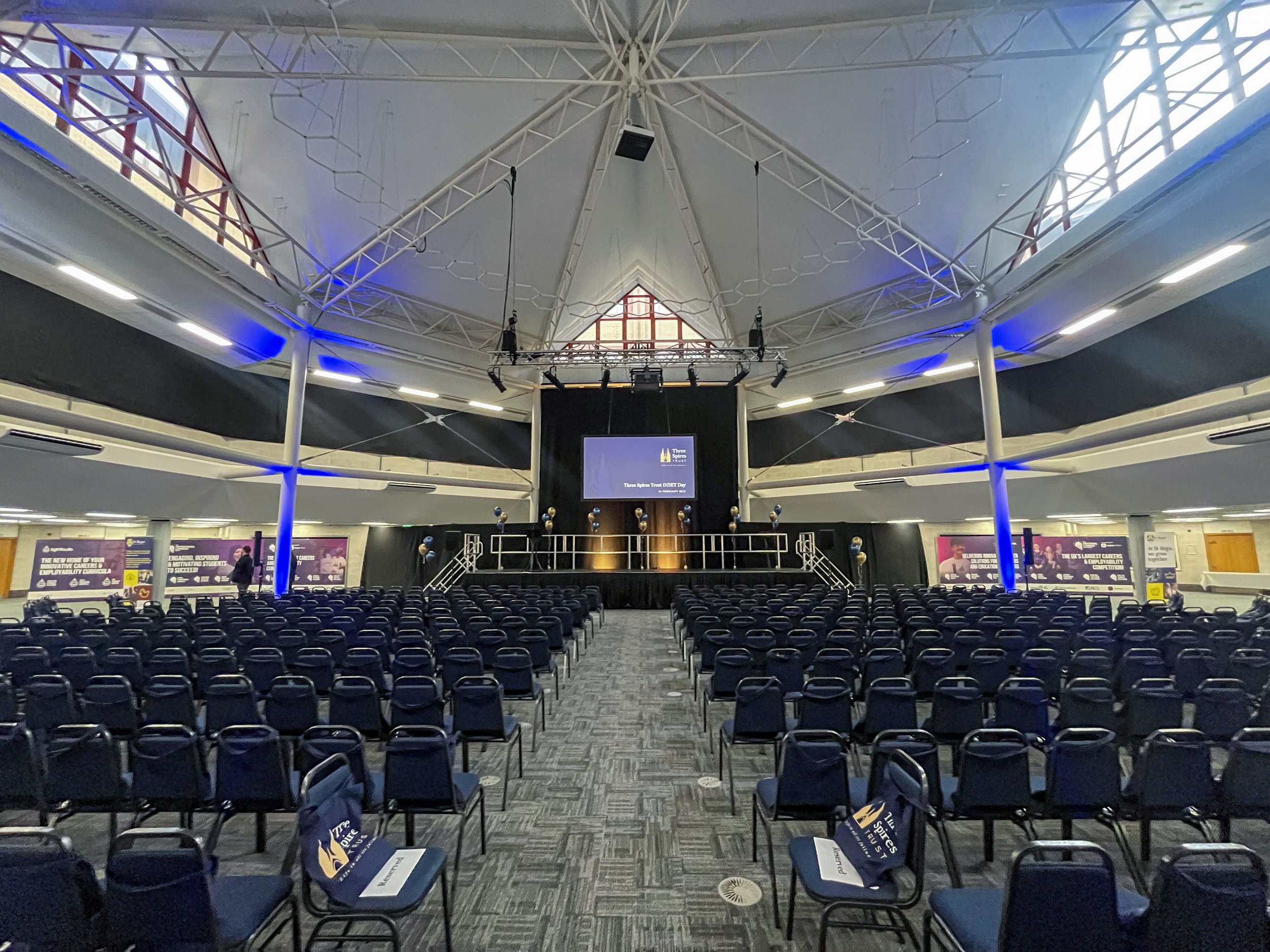 An empty conference or event hall with rows of chairs facing a stage with a large screen and lighting equipment, under a high, geometric ceiling.