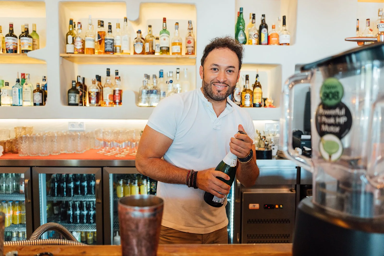 A man with dark hair and a beard smiling and holding a bottle of sparkling wine in a bar with shelves of liquor bottles in the background.