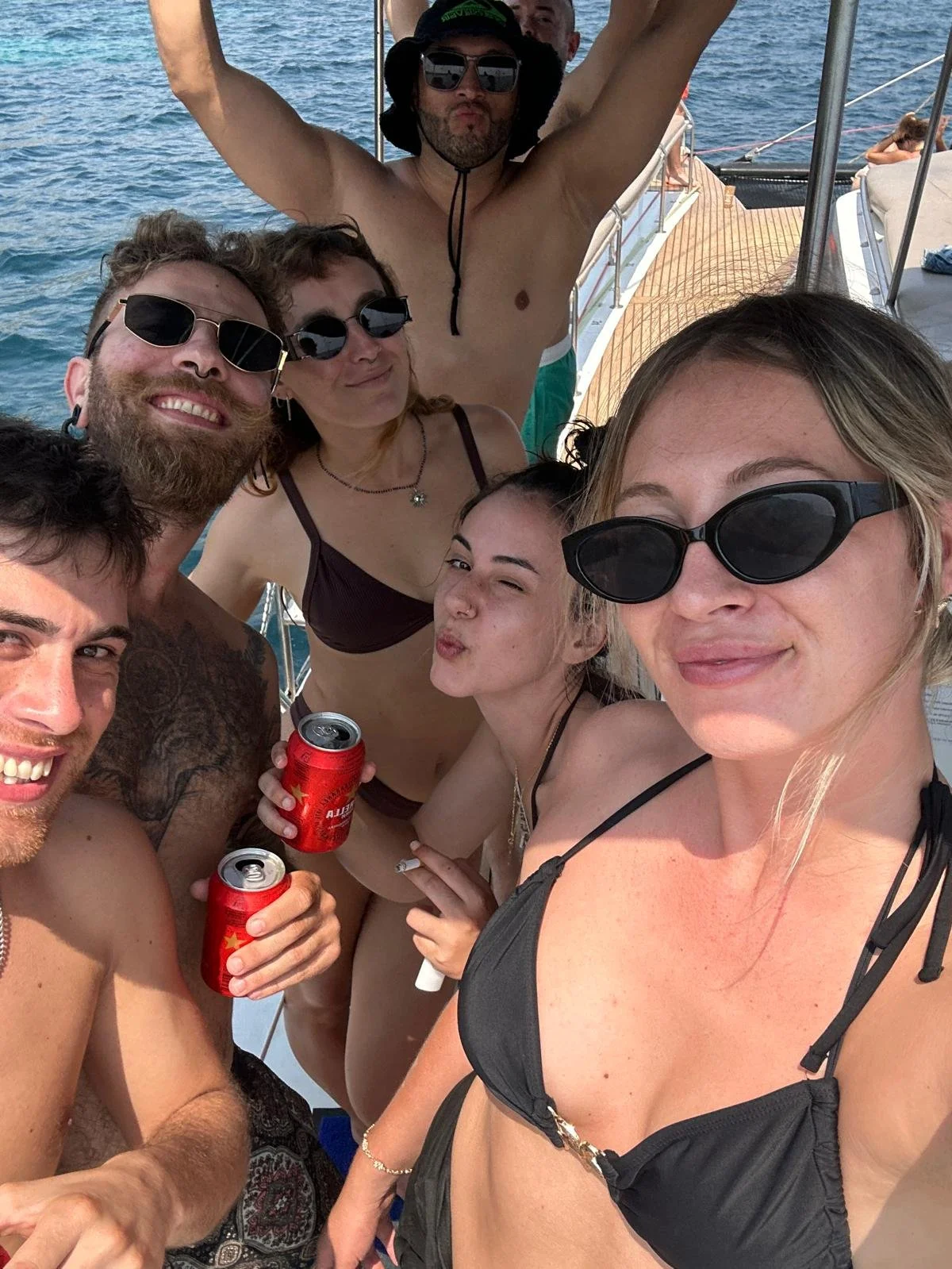 Group of friends taking a selfie on a boat, wearing swimsuits and sunglasses, with the ocean in the background.