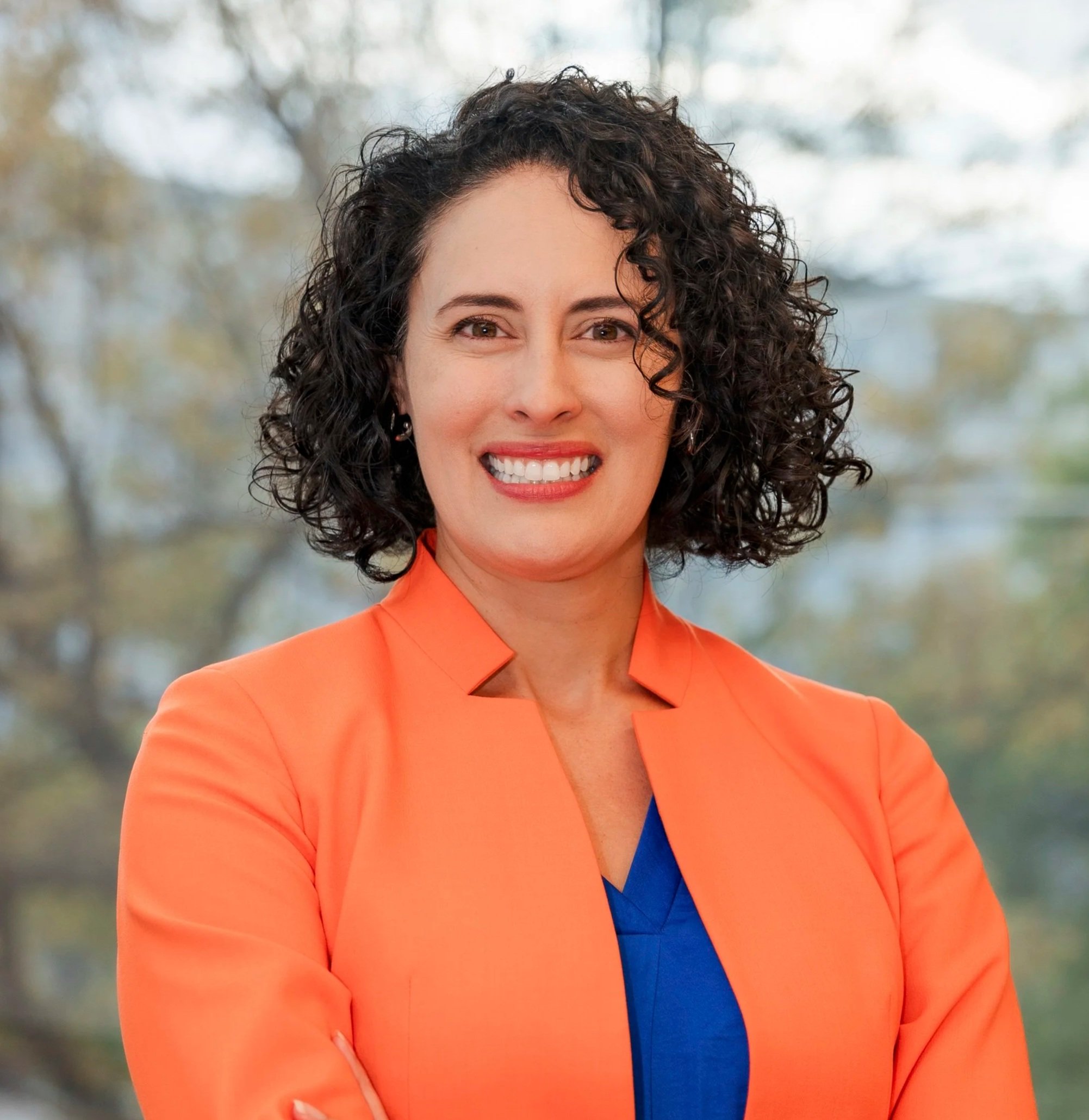 Professional woman with curly dark hair smiling, wearing an orange blazer over a blue top, standing outdoors with blurred trees in the background.