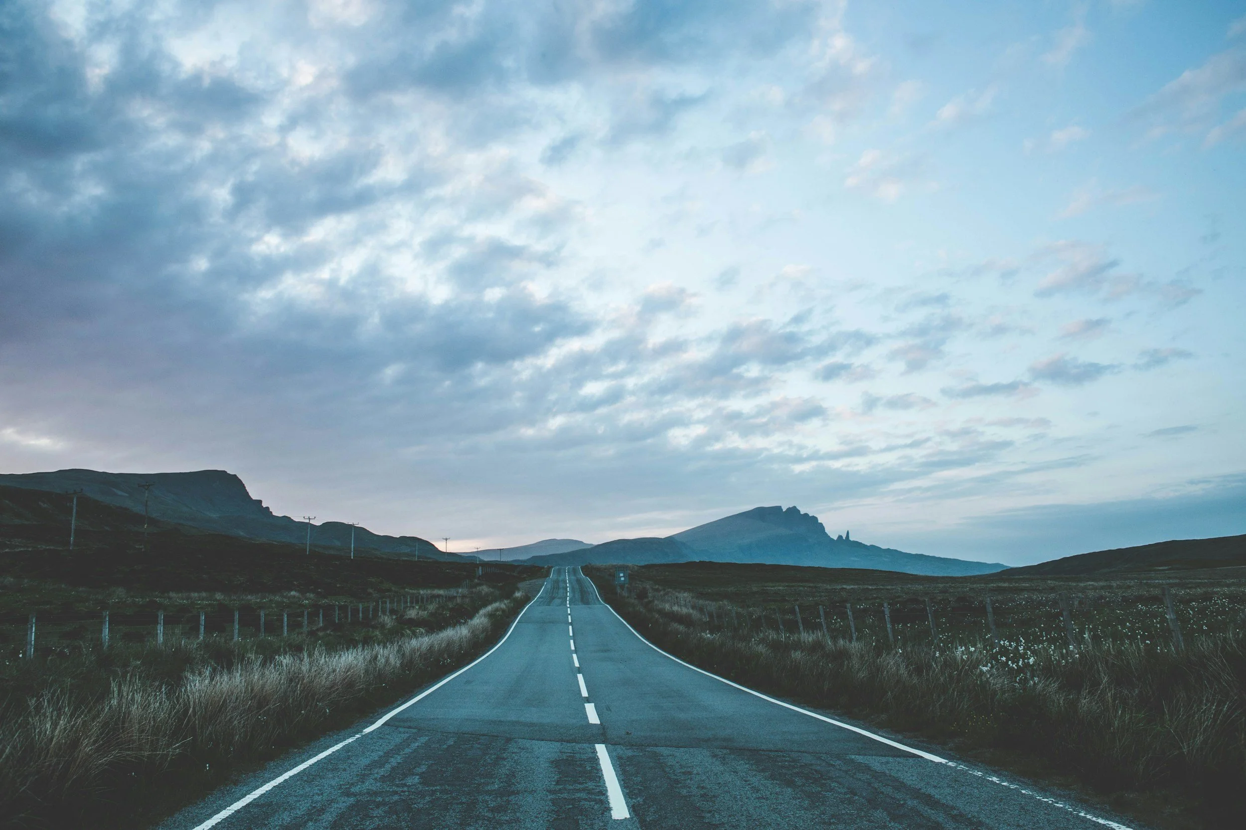 A straight empty road running through a rural landscape with mountains in the background, under a cloudy sky.