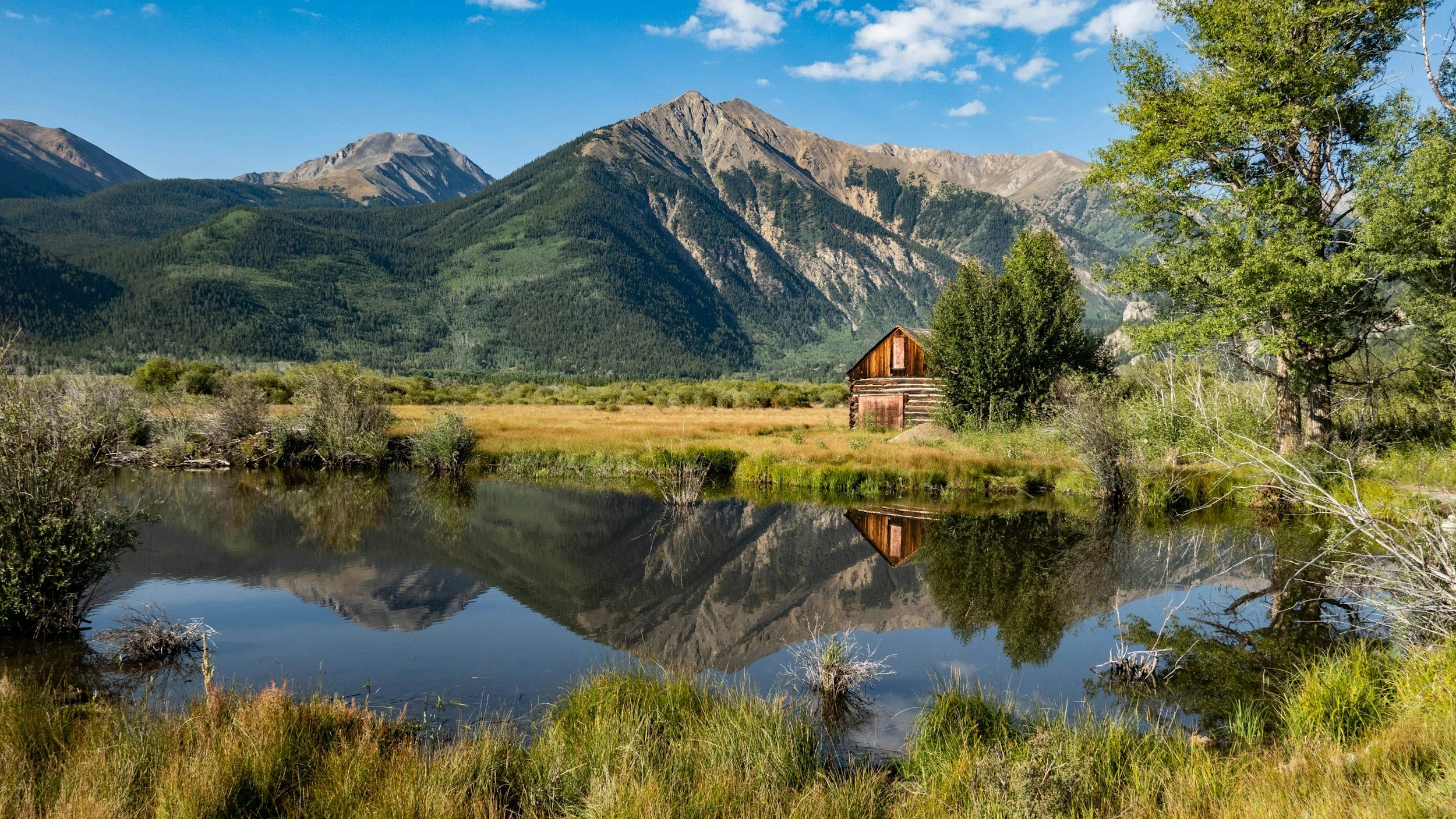 Mountain landscape with a small wooden cabin near the water, surrounded by green trees and shrubs, with mountains in the background and a clear blue sky.